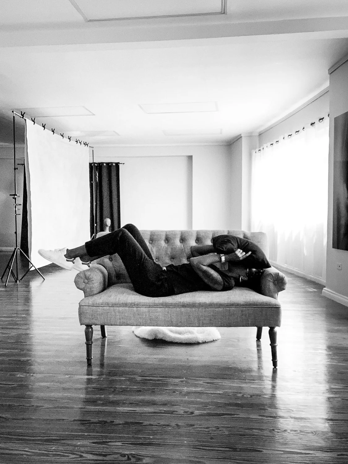 Person lying on a vintage-style sofa in a room with hardwood floors, curtains, and photography equipment.