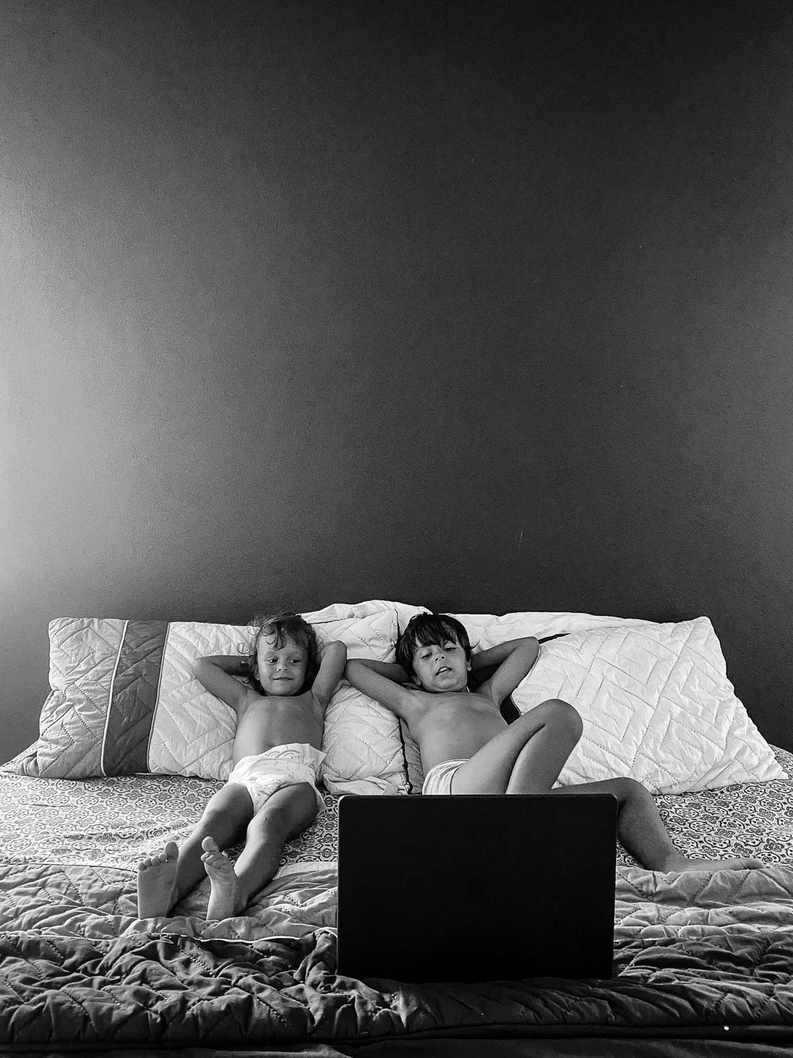 Two young children lying on a bed with pillows, watching a laptop.