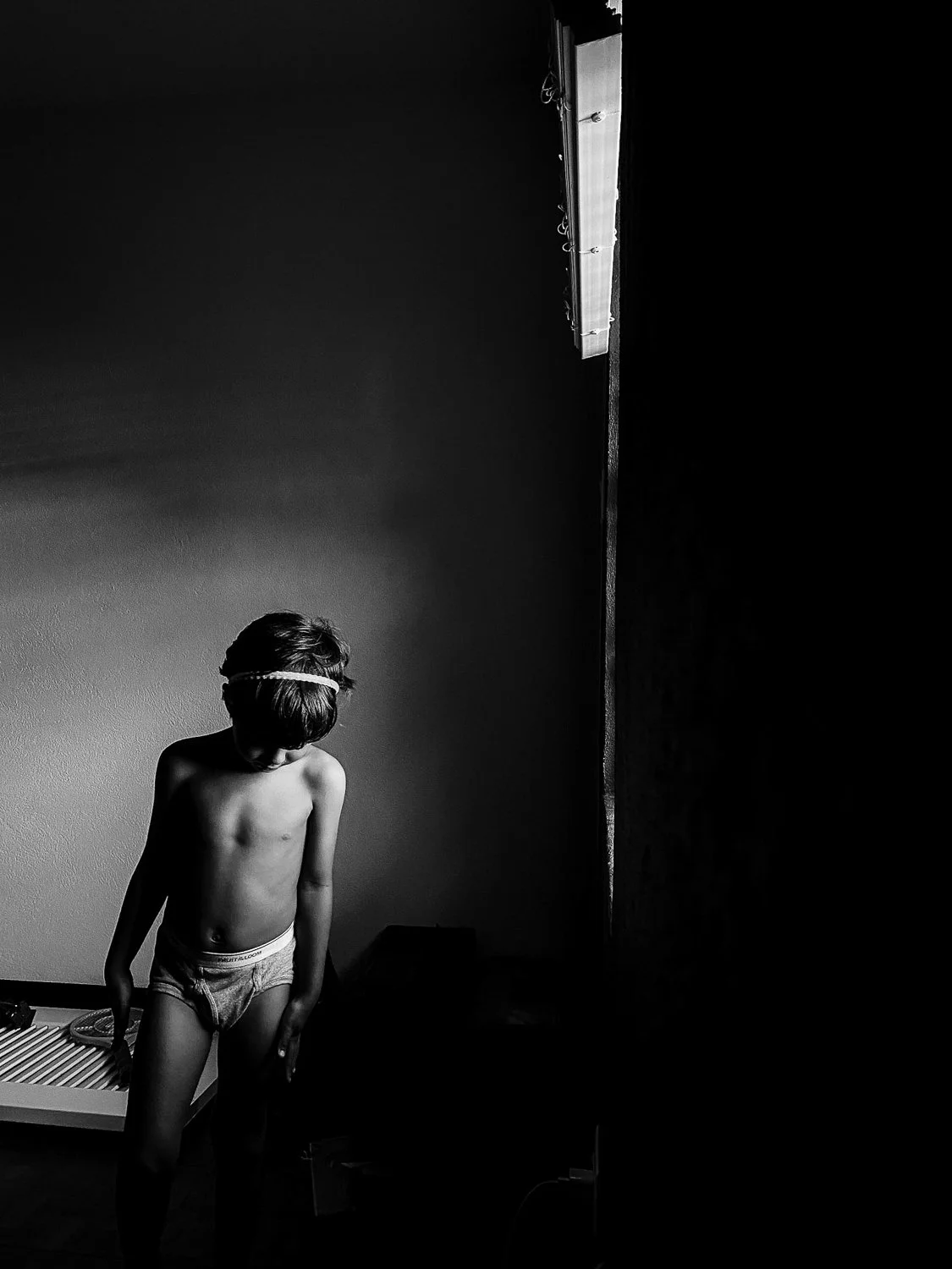 A young boy with a headband, shirtless, wearing underwear, standing in a dimly lit room, looking downward with one hand hanging down, near a bed and a wall with a fluorescent light fixture.