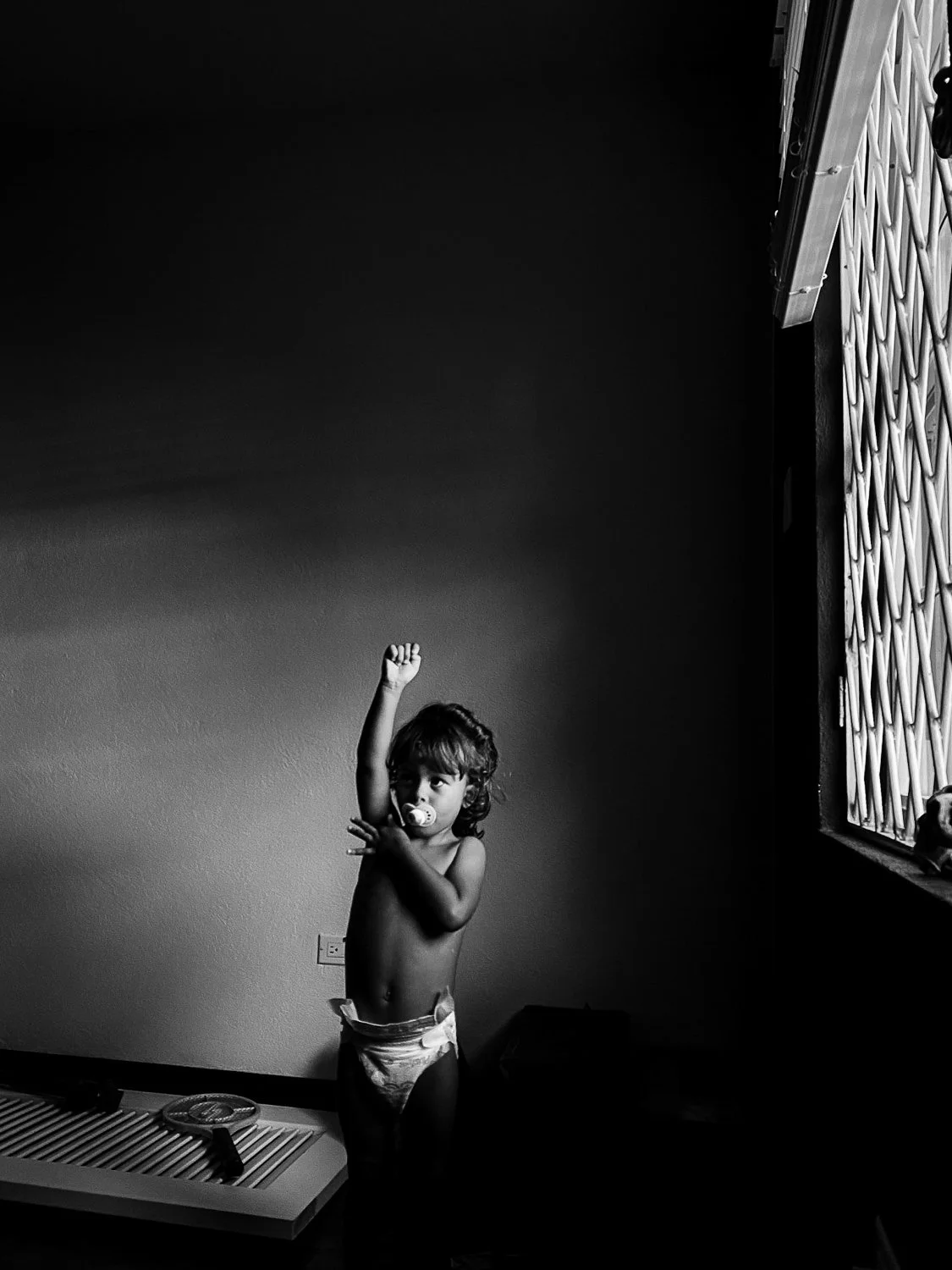 A young child wearing only a diaper, with a pacifier in mouth, standing in a room near a window, raising one arm and looking towards the camera.