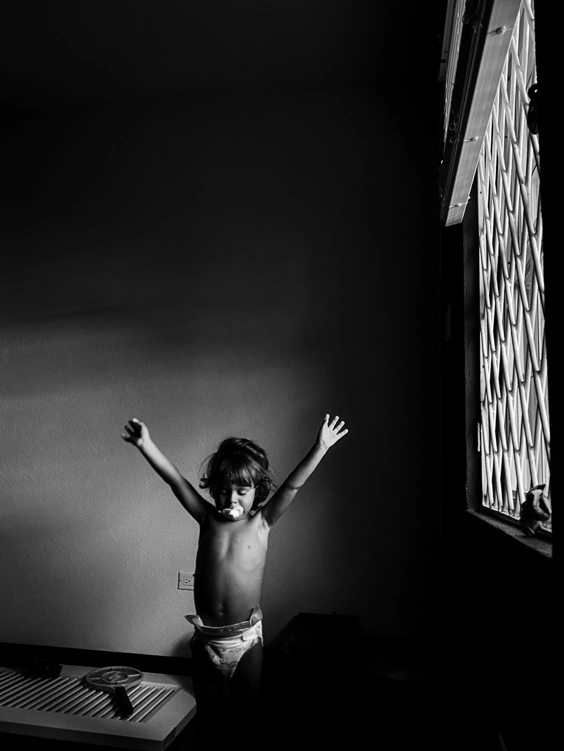 A young child with a pacifier in their mouth standing shirtless with arms raised in a sunlit room with a barred window on the right side.