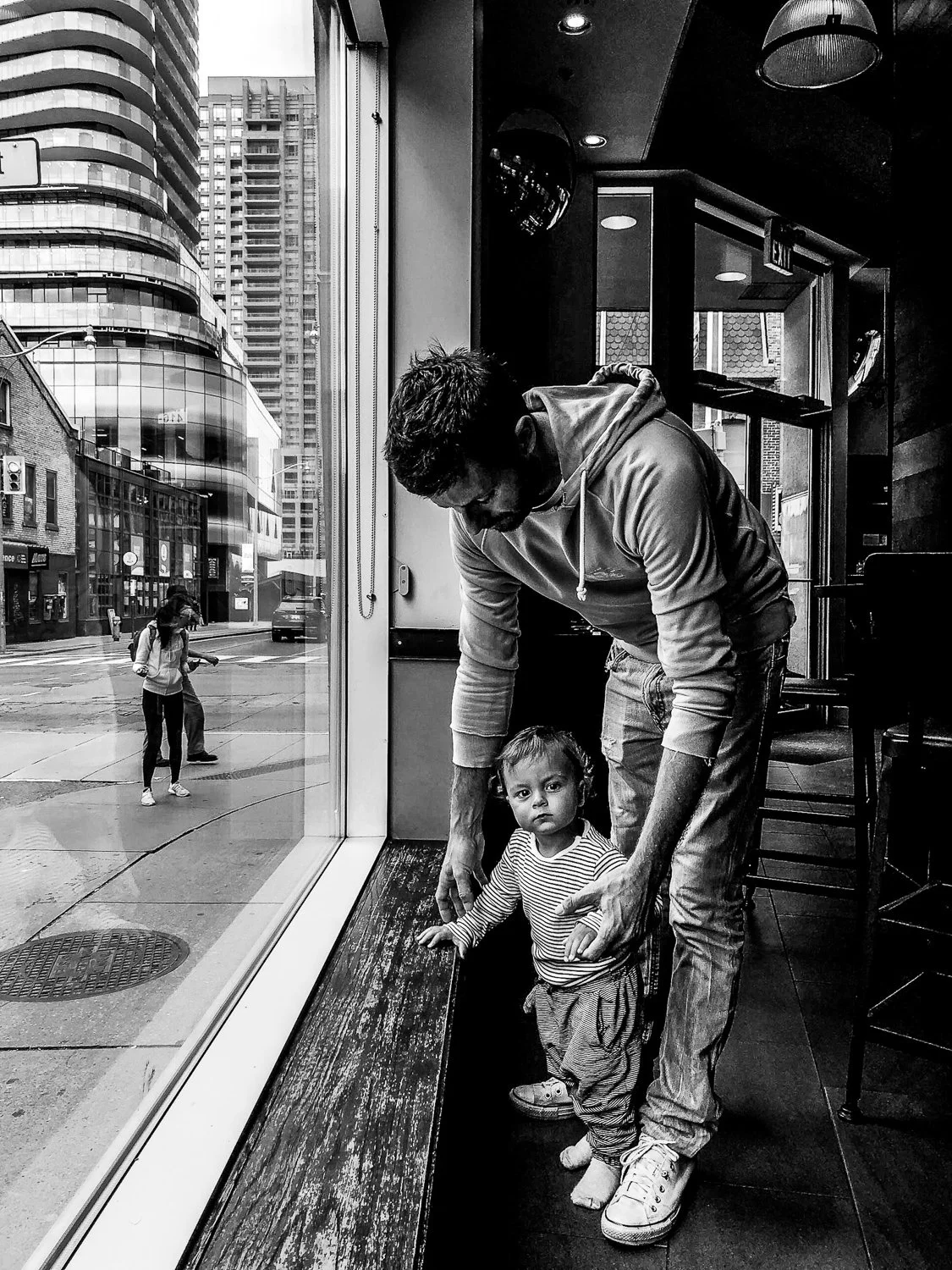 A man and young girl standing inside a cafe near a large window, with the city street and pedestrians visible outside.