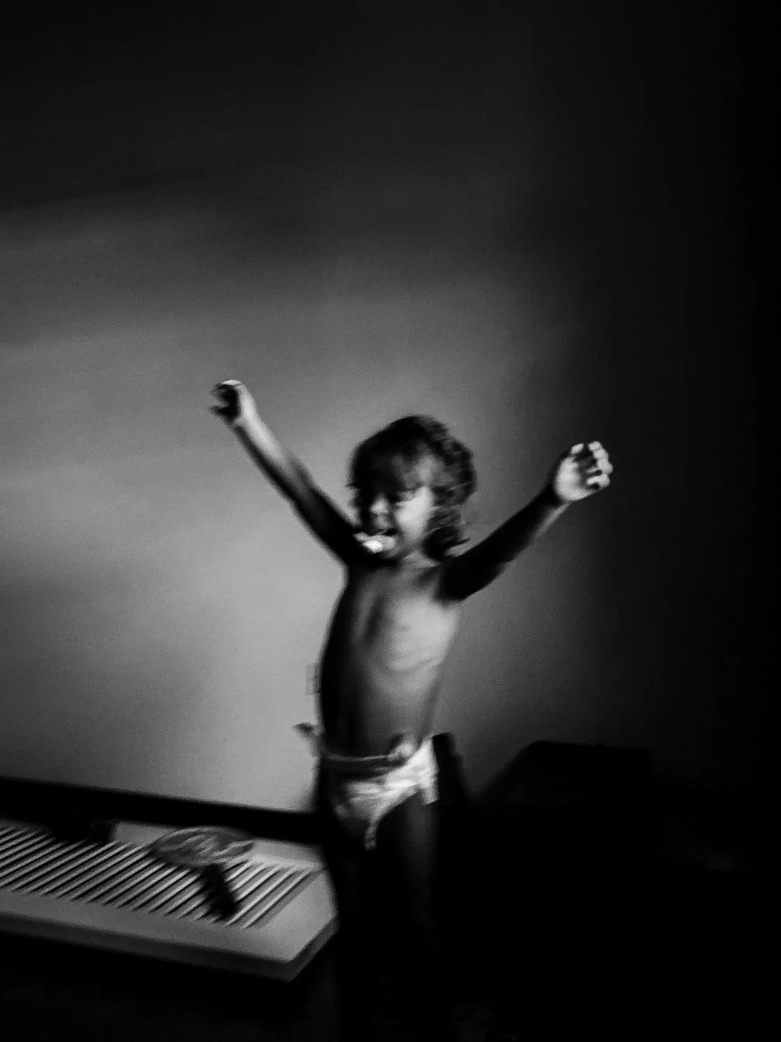 A black and white photo of a shirtless child with arms raised and a wide-open mouth, standing next to a small appliance on a flat surface against a plain background.