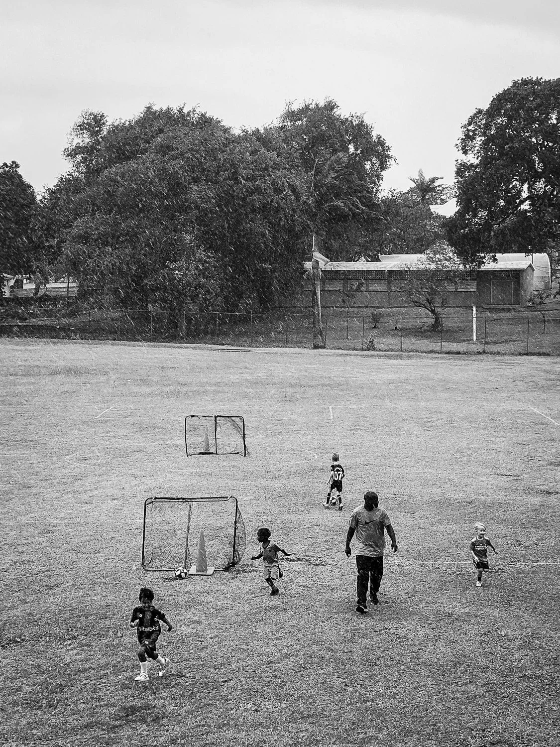 Children playing soccer on a grassy field with a coach or adult, with soccer goals and cones, and trees in the background in black and white.