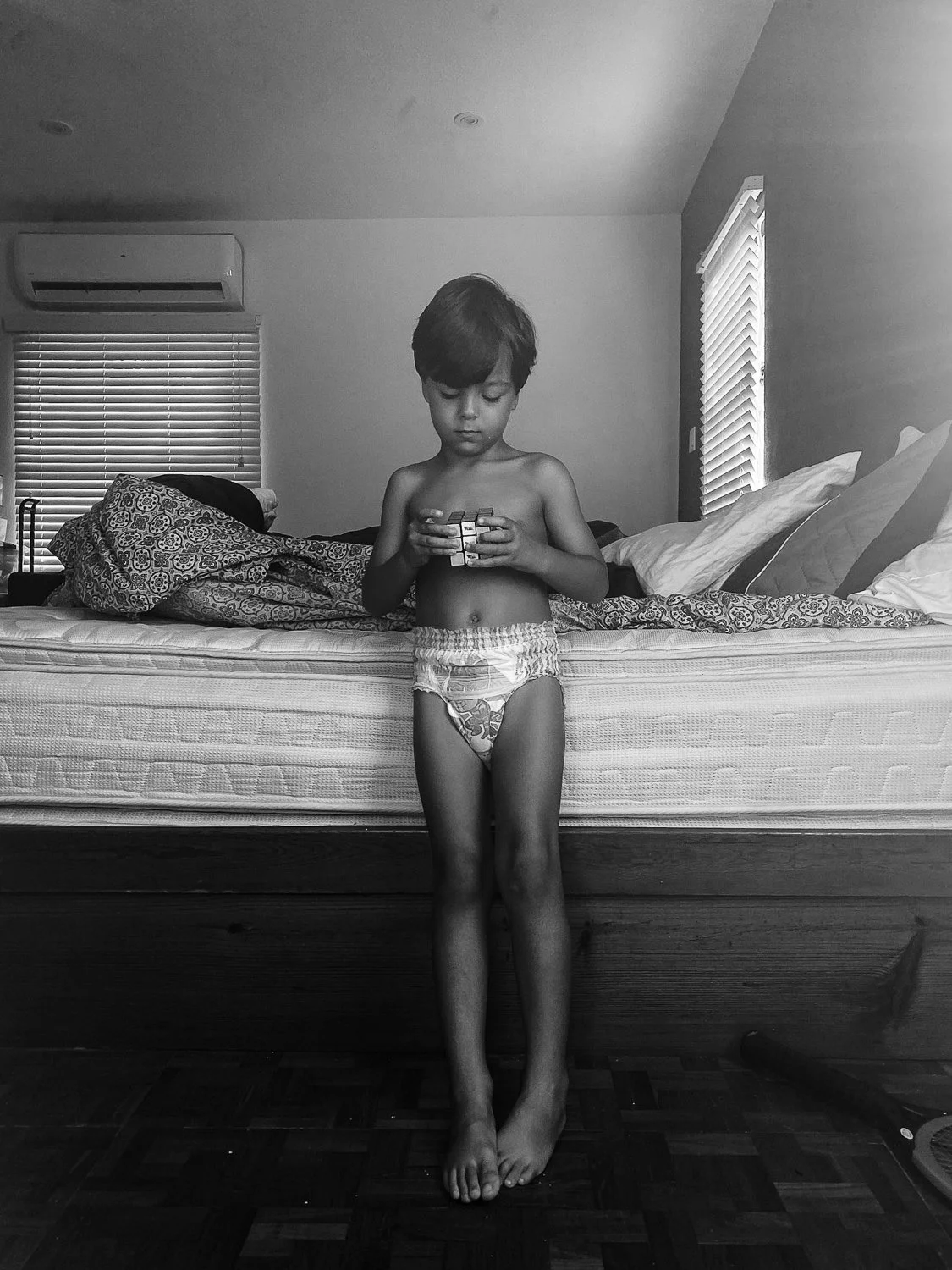 A young boy in underwear stands barefoot in front of a bed, playing with a Rubik's Cube in a bedroom with blinds on the windows.