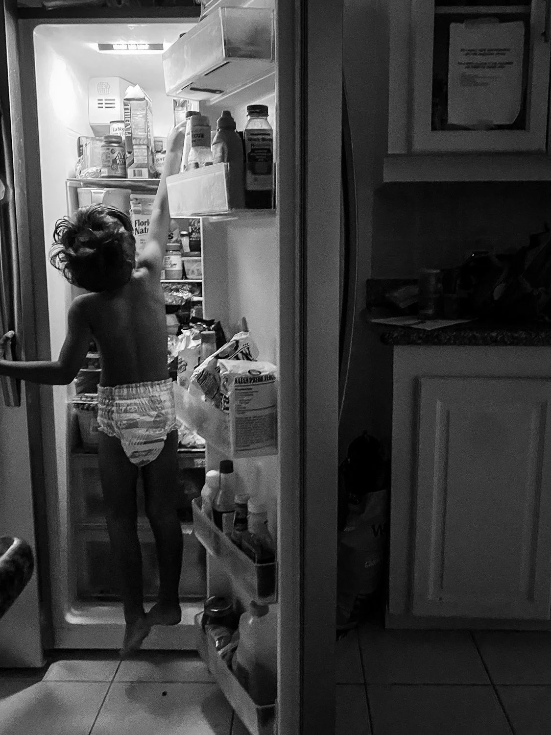 A young child with curly hair standing inside an open refrigerator, reaching for a bottle on the top shelf, wearing only a diaper.