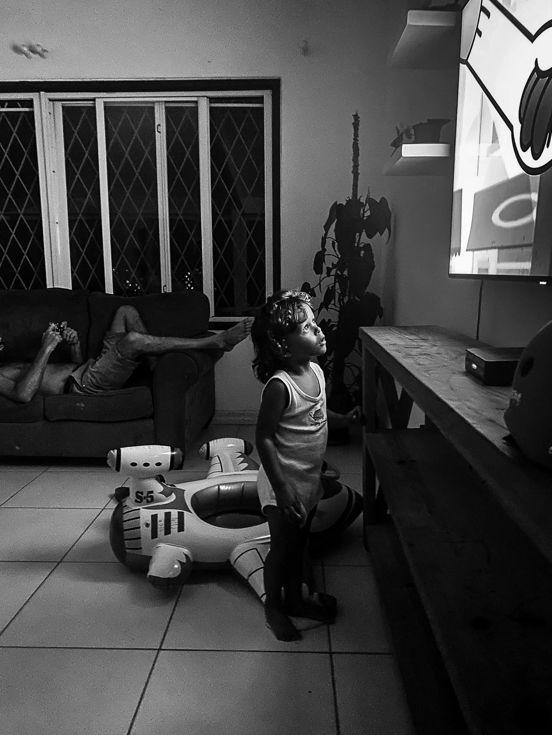 A young girl standing on the floor watching TV in a living room at night, with two children lounging on a sofa in the background, and an inflatable airplane on the floor behind her.