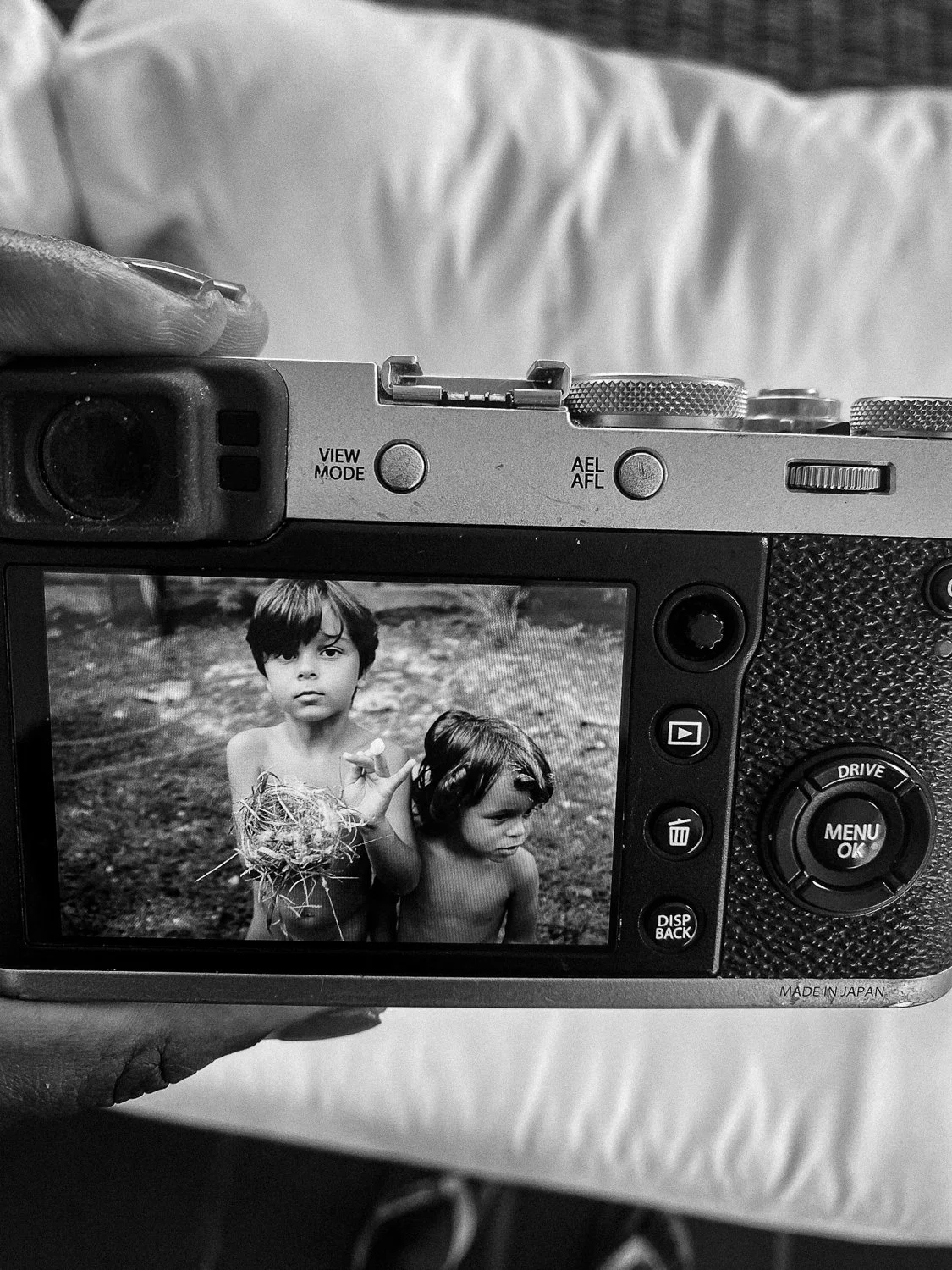 A person holding a camera shows a black-and-white photo of two young boys, one holding a ball of tangled string, outdoors on the camera's screen.
