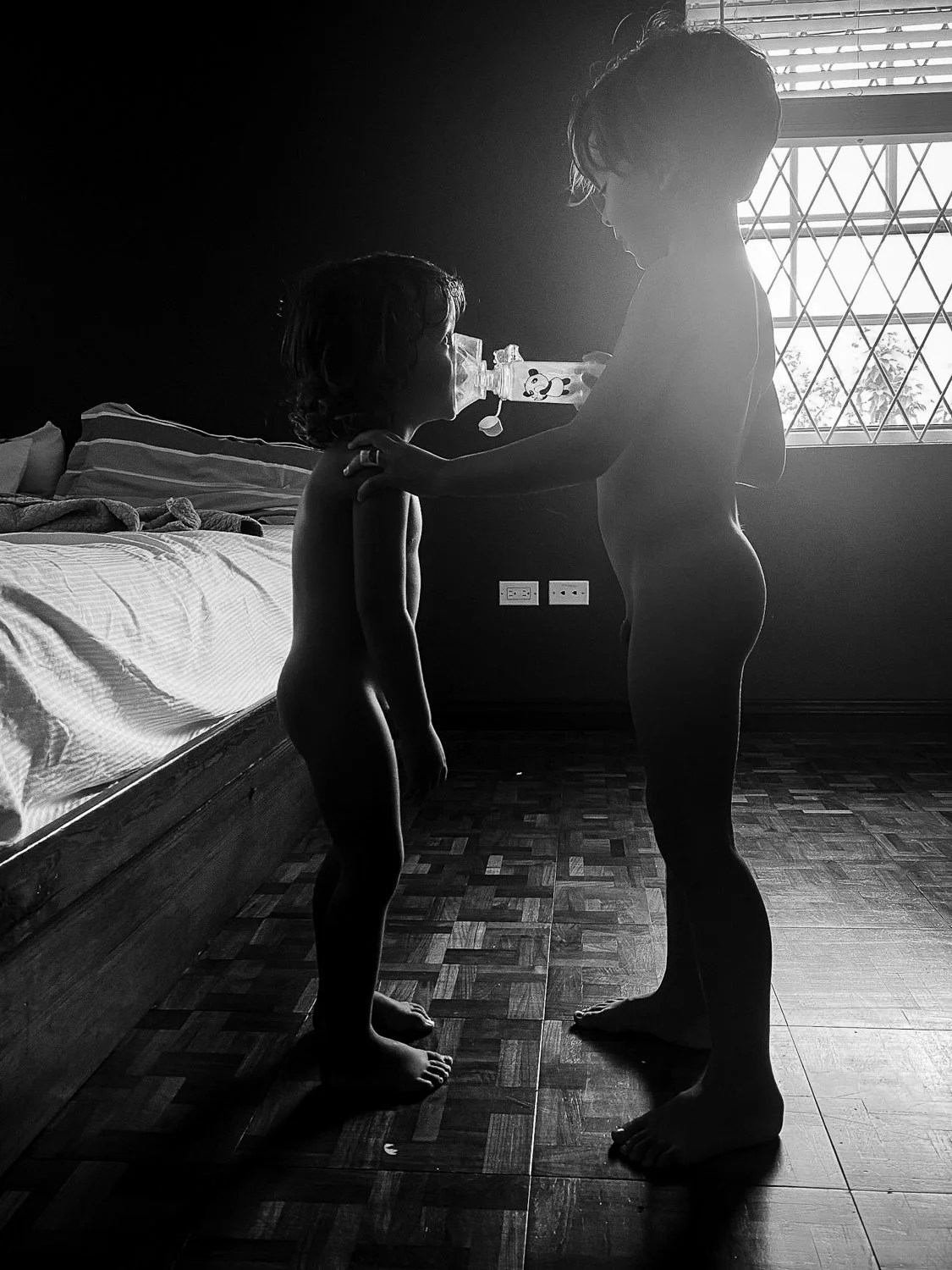 Two children, one older and one younger, standing face to face in a dimly lit room. The older child is holding a water bottle to the younger child's mouth, who is drinking. The light from a window behind them creates a silhouette effect.