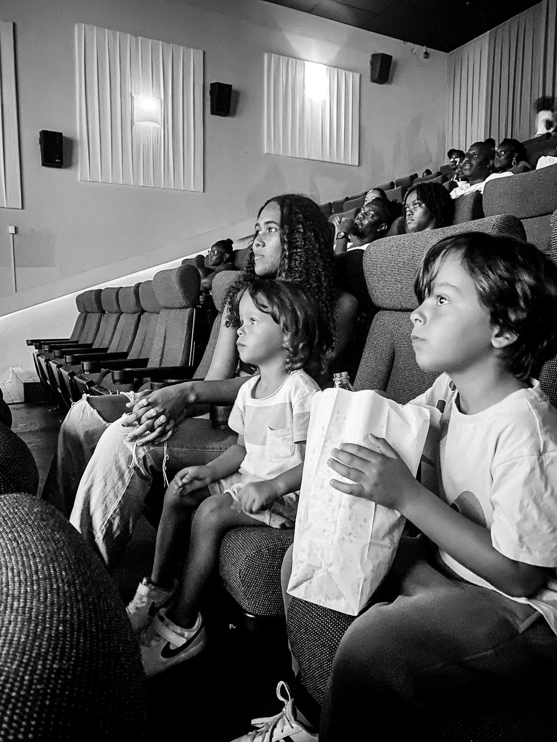 People watching a movie or presentation in a theater, with children and adults seated in dimly lit auditorium