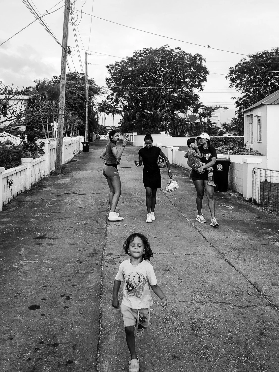 A black and white photo of a neighborhood street with five people. A young girl is running towards the camera in the foreground, while four women and a small child are walking and talking in the background.