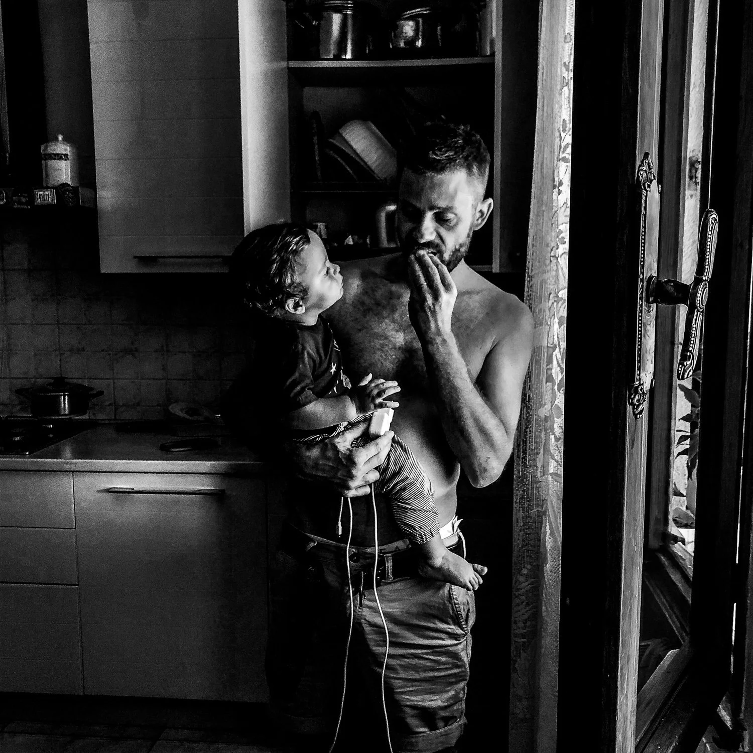 A shirtless man holding a young child in a kitchen near an open door, with the man touching his lips. The child is looking at the man and sitting on his arm, wearing patterned pants.