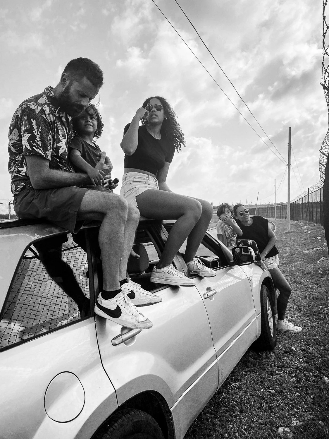 A group of five people sitting on and around a car parked near a barbed wire fence on a partly cloudy day.