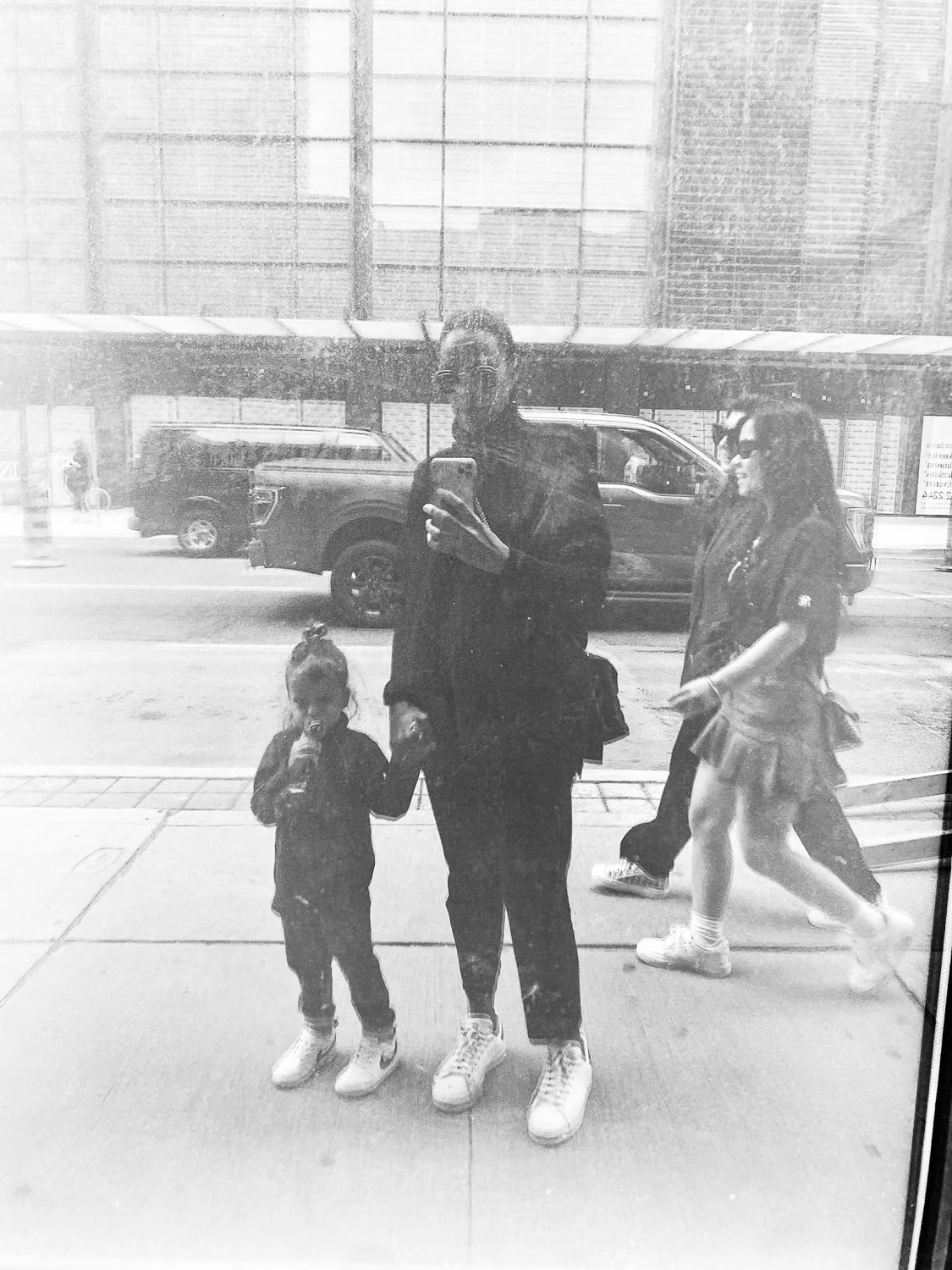 Reflected image of a man with glasses holding a child's hand, standing on a city sidewalk, with pedestrians and cars in the background, seen through a glass window.