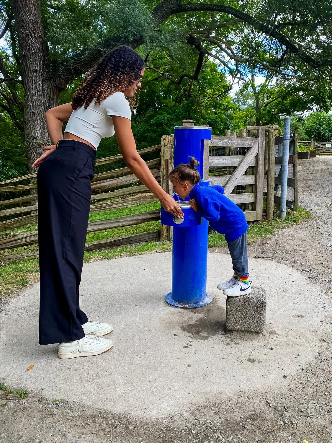 A woman with curly hair and wearing black pants and a white crop top helps a young girl with brown hair in a ponytail drink water from a public drinking fountain at a park. The girl is standing on a small concrete block and leaning over to drink from