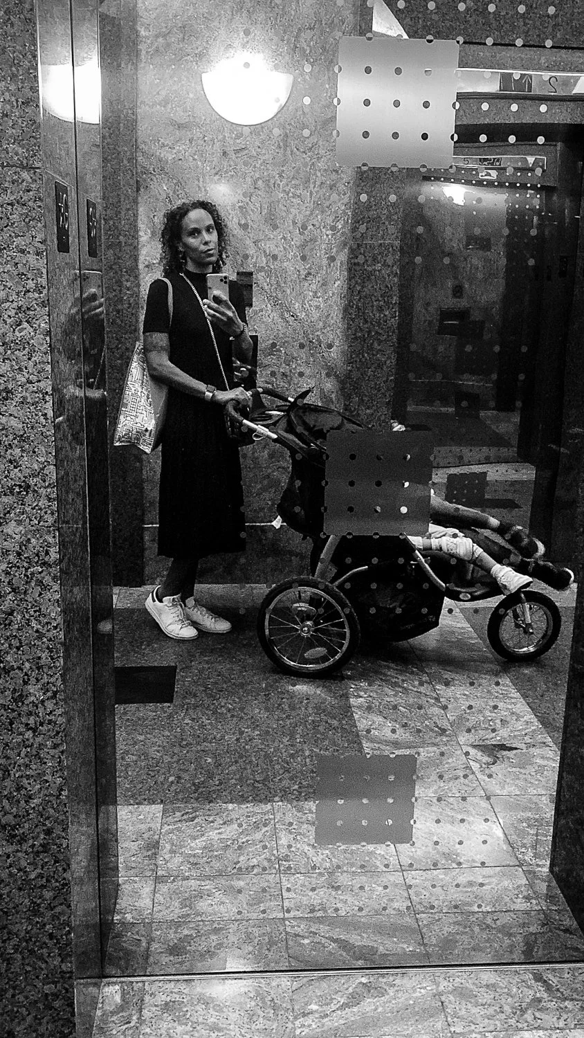 A woman taking a selfie in an elevator mirror, pushing a stroller with a child inside, in a building with marble walls and floors.