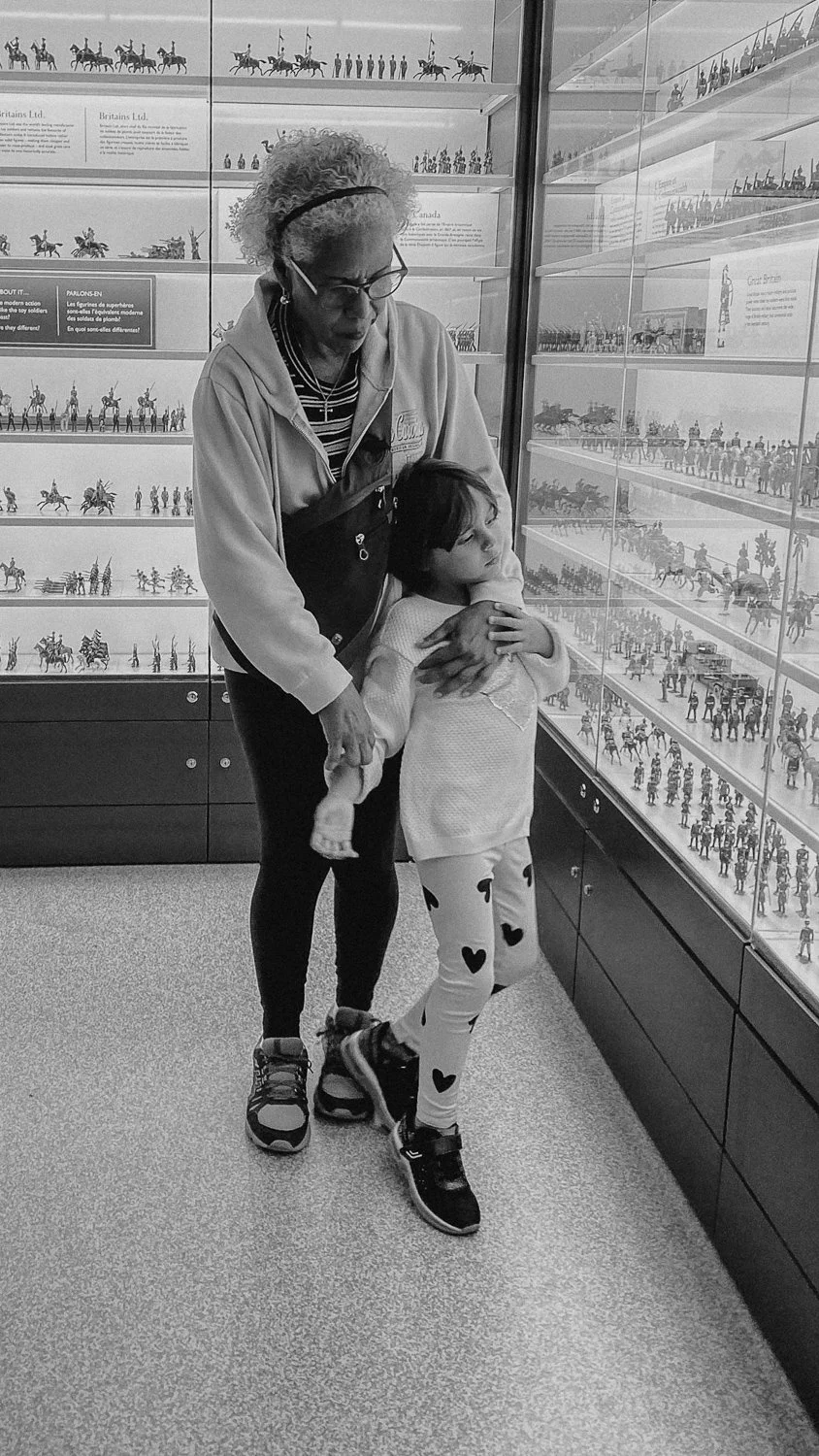 A woman and a young girl standing in front of glass display cases filled with miniature figurines of historical soldiers and horses. The woman is wearing glasses, an athletic jacket, and is gently holding the girl, who is leaning into her with a thou
