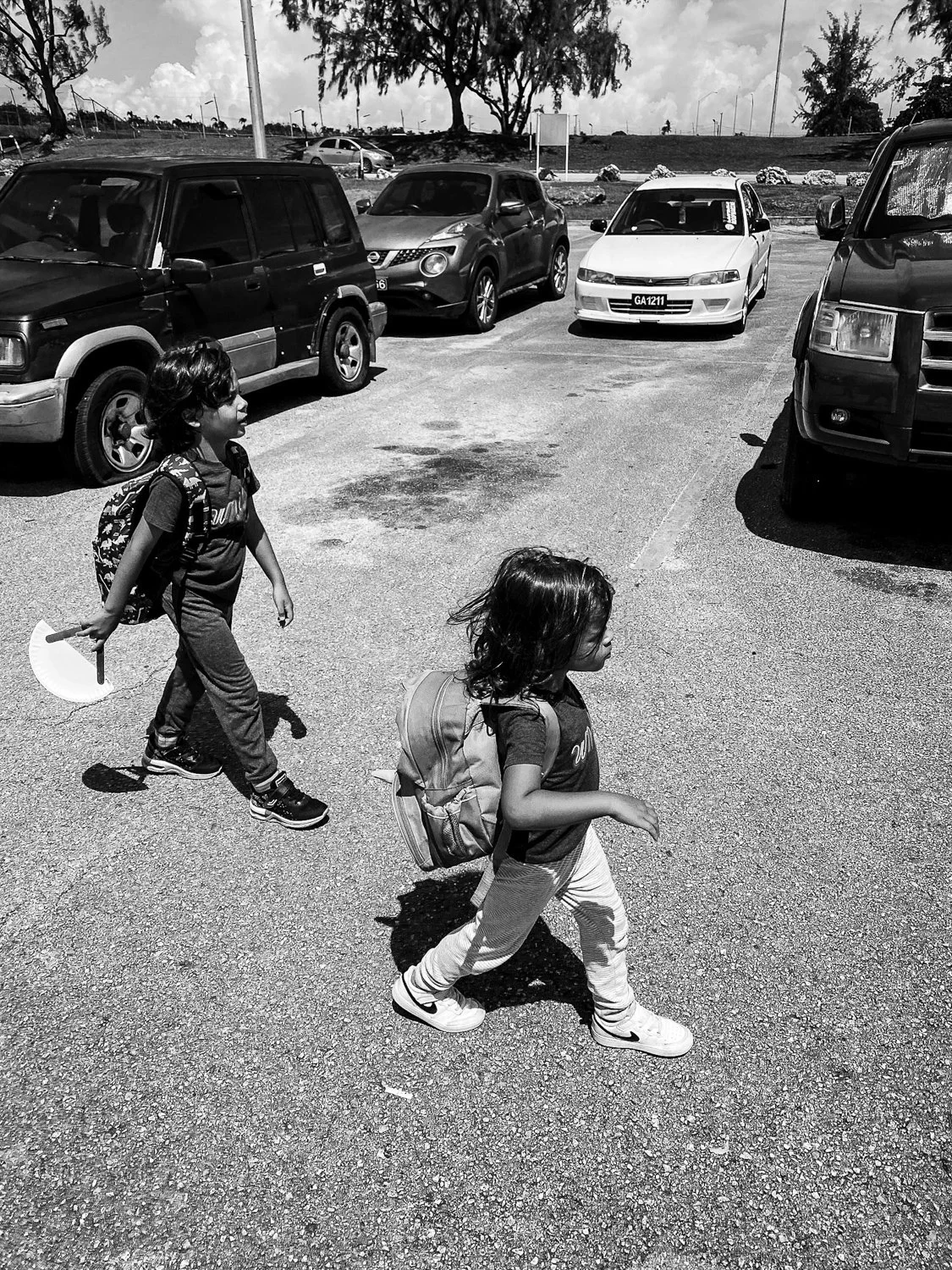Two children walking across a parking lot with backpacks, surrounded by parked cars and trees.