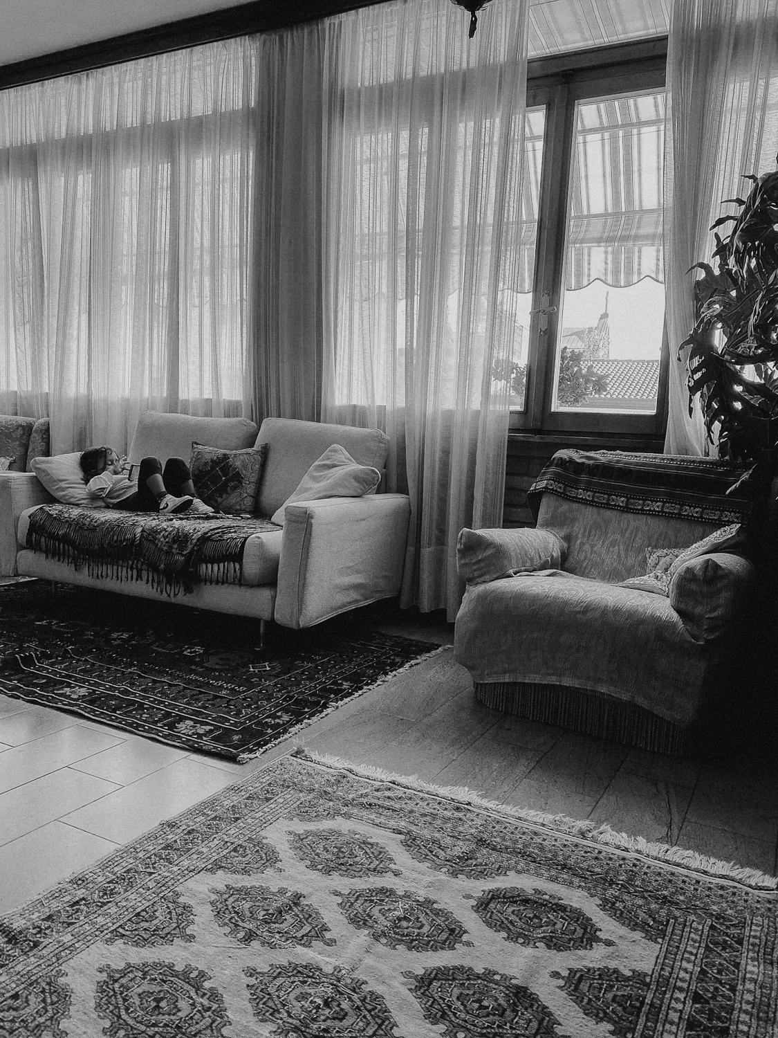 Living room with large windows covered by sheer curtains, featuring a young girl lying on a sofa, pillows, and a patterned rug on the floor. An armchair with a cushion is nearby, and a potted plant is in the corner.
