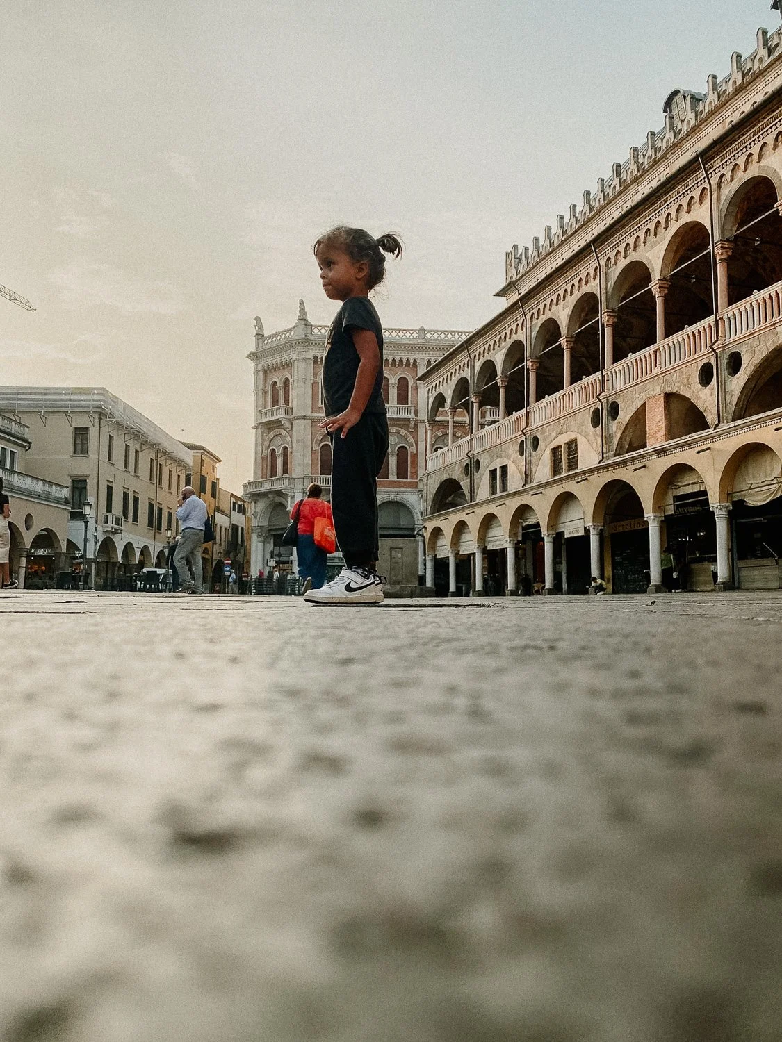 A young girl standing on a cobblestone street in front of historic European-style buildings at sunset.