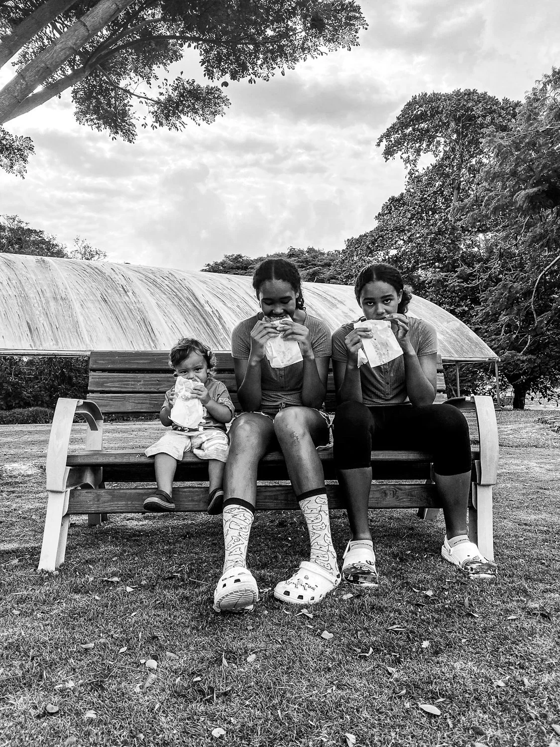 Three children sitting on a park bench, eating fast food, with trees and an arched structure in the background.