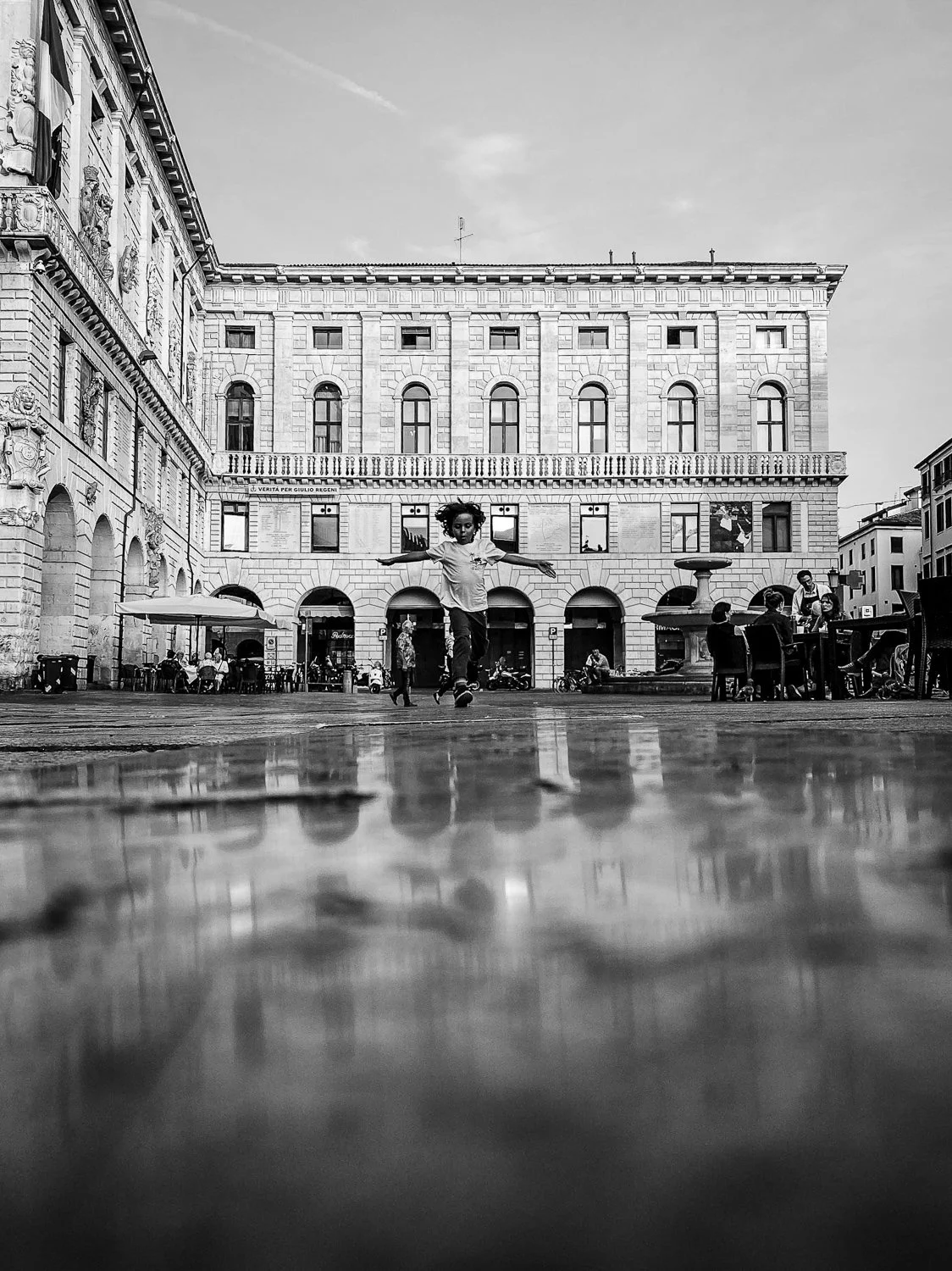 A child jumping in a public square with a large historic building and outdoor seating in the background, reflected in a wet surface in the foreground.