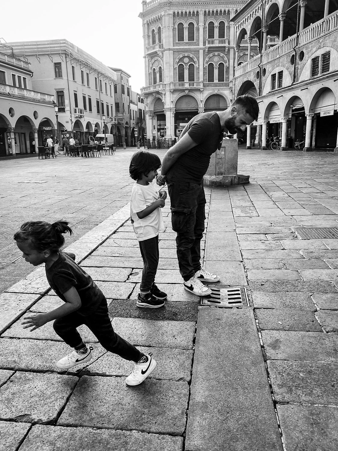 A man with two children in a European city square. One child is jumping, while the other child and the man stand on a drain cover, with historic buildings in the background.