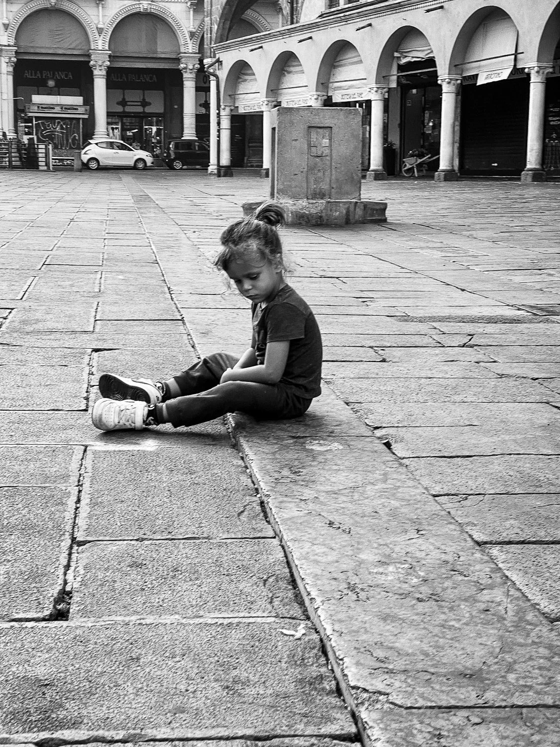 A young girl sitting on a sidewalk, looking down, in a plaza with historic arches and buildings in the background.