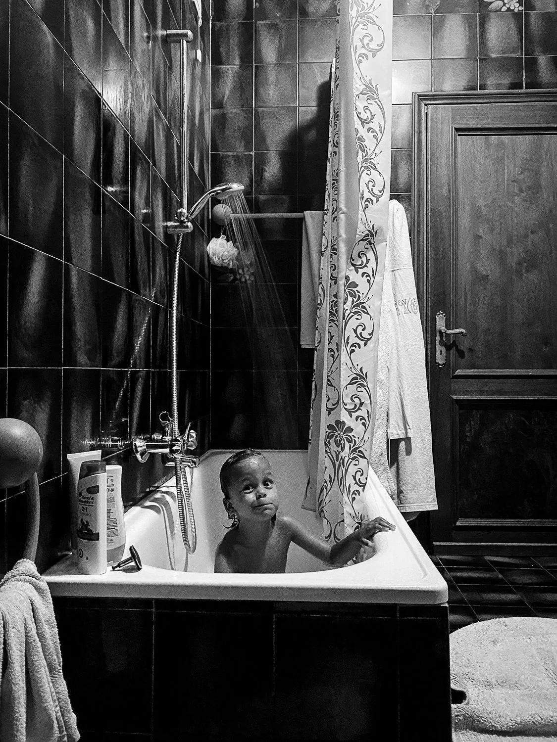 A young child sitting in a bathtub filled with water in a bathroom with dark tiled walls, a shower head, a curtain with floral patterns, and a towel hanging on a hook.