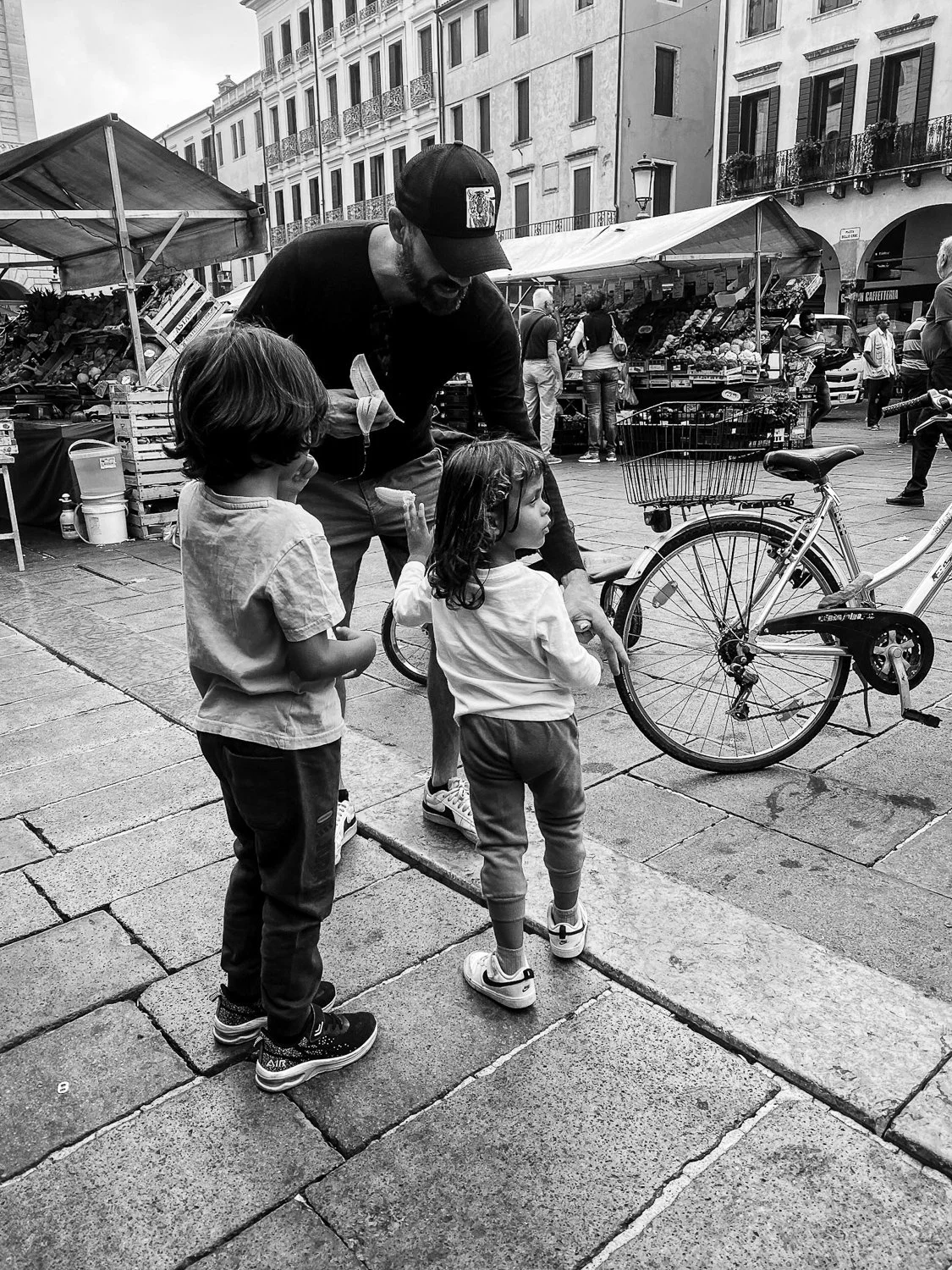 A man with a beard wearing a black cap interacts with three young children on a city street, with market stalls and buildings in the background.