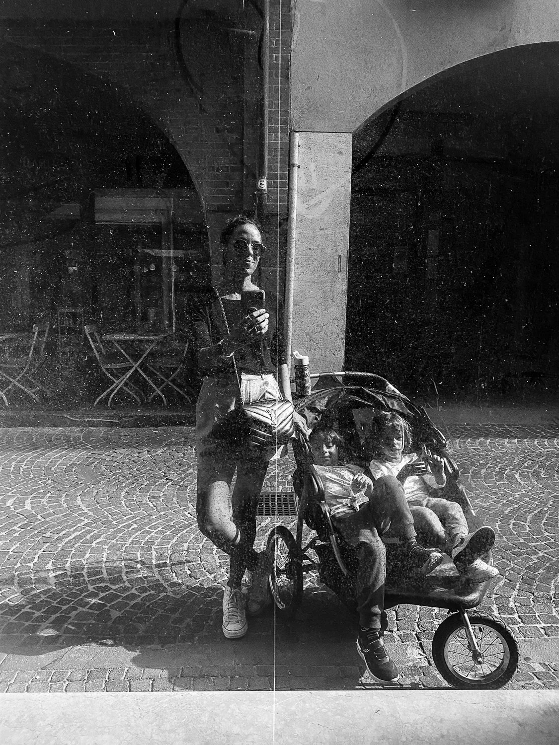 Reflection of a woman taking a selfie in a glass window, with two children seated in a stroller in front of her, on a cobblestone street.