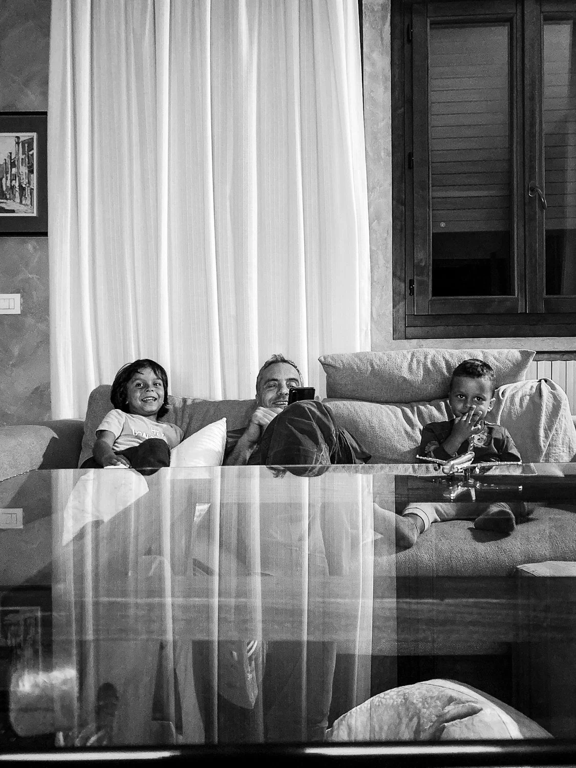 A family of three sitting on a couch in a living room, smiling and looking towards the camera, with a glass coffee table in the foreground.