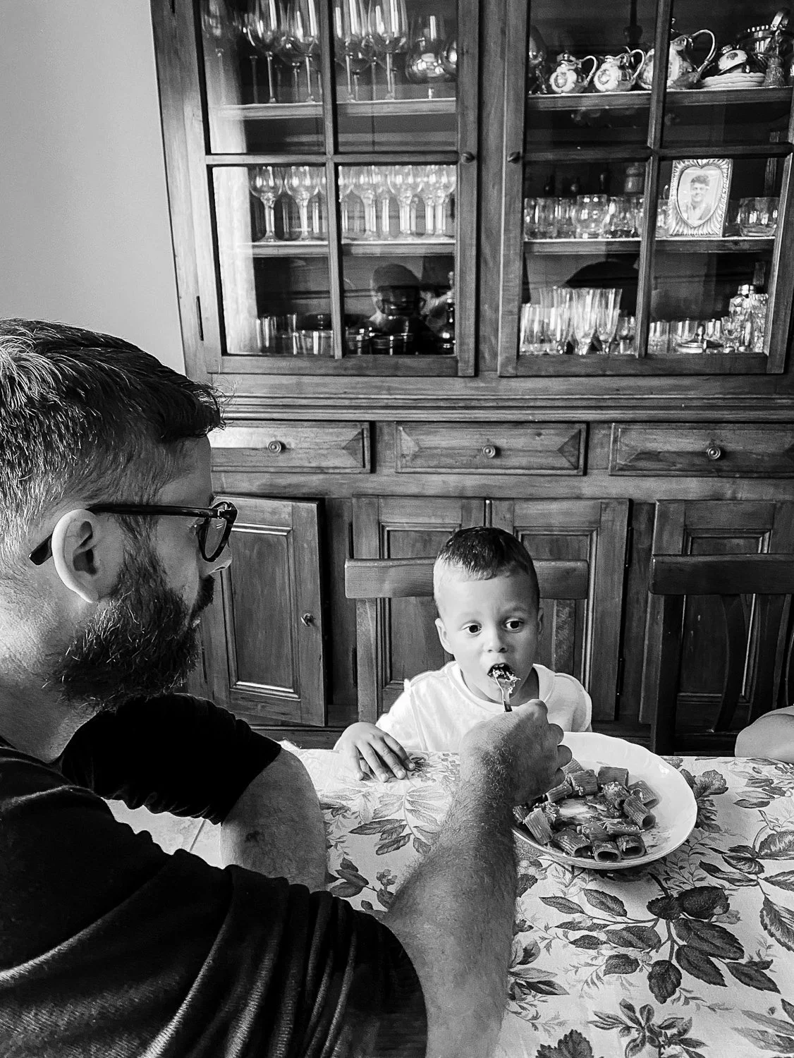 A man with glasses and a beard feeding a young boy with a fork at a dining table. The boy has a plate of food in front of him. A wooden china cabinet with glass doors filled with glassware and decorative items is in the background.