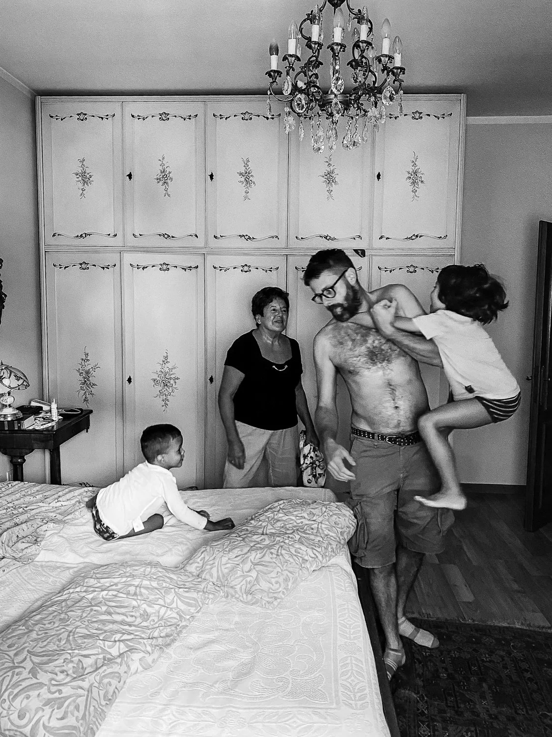 A family in a bedroom with vintage furniture and ceiling chandelier. A shirtless man with glasses holds a young girl in a white shirt and striped shorts. A woman and a young boy are on the bed.