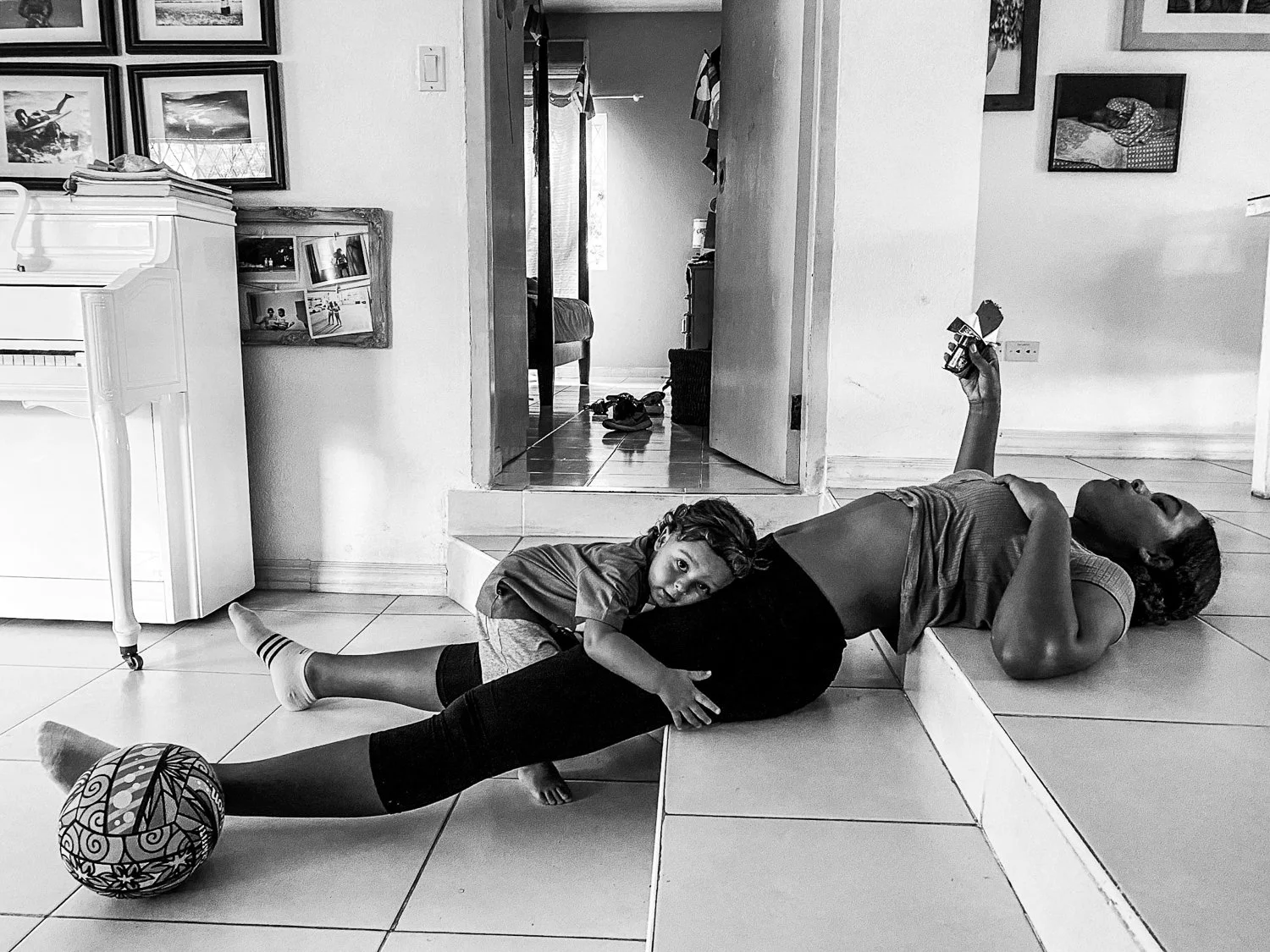 A girl lying on the floor playing with a boy who is hugging her from behind. The girl is lying on her back with her head turned to the side, holding a phone in her right hand. The boy is lying on his stomach with his head resting on the girl's stomac