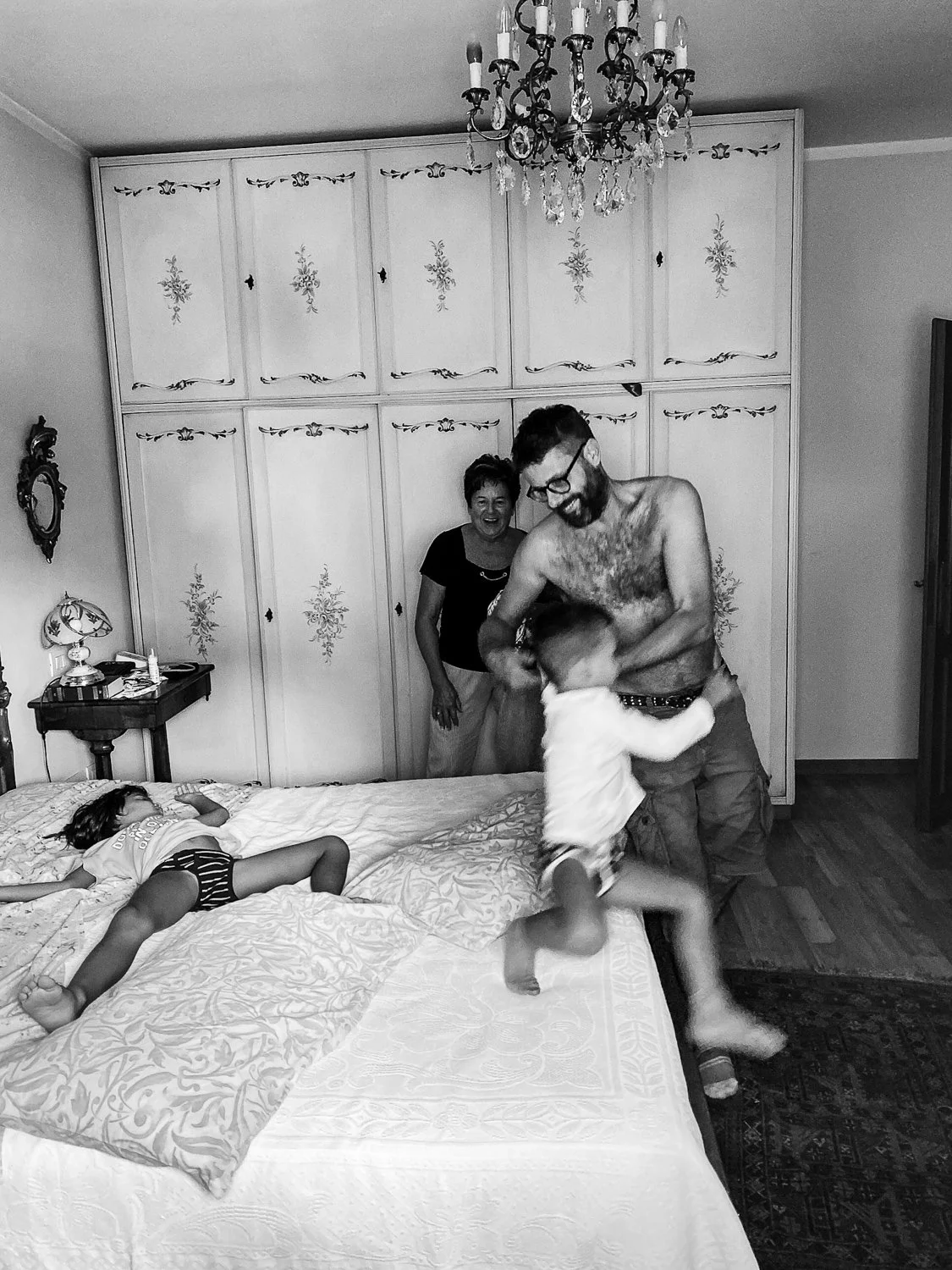 A man playing with a child on a bed while a woman and another girl watch in a bedroom with a chandelier and decorated cabinets.