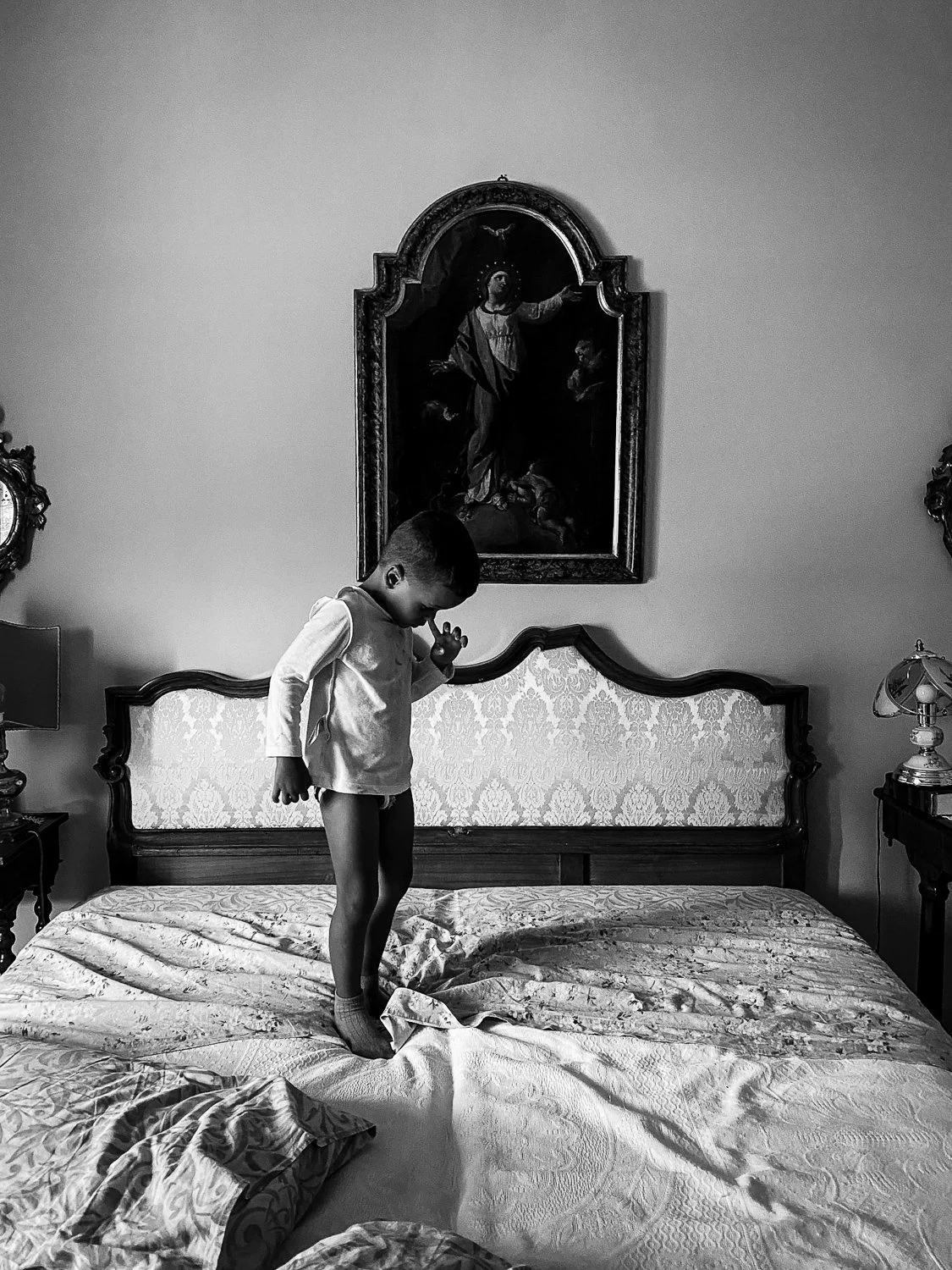 A boy standing on a bed in a bedroom, with a religious painting hanging above the headboard, and two bedside lamps on either side of the bed.