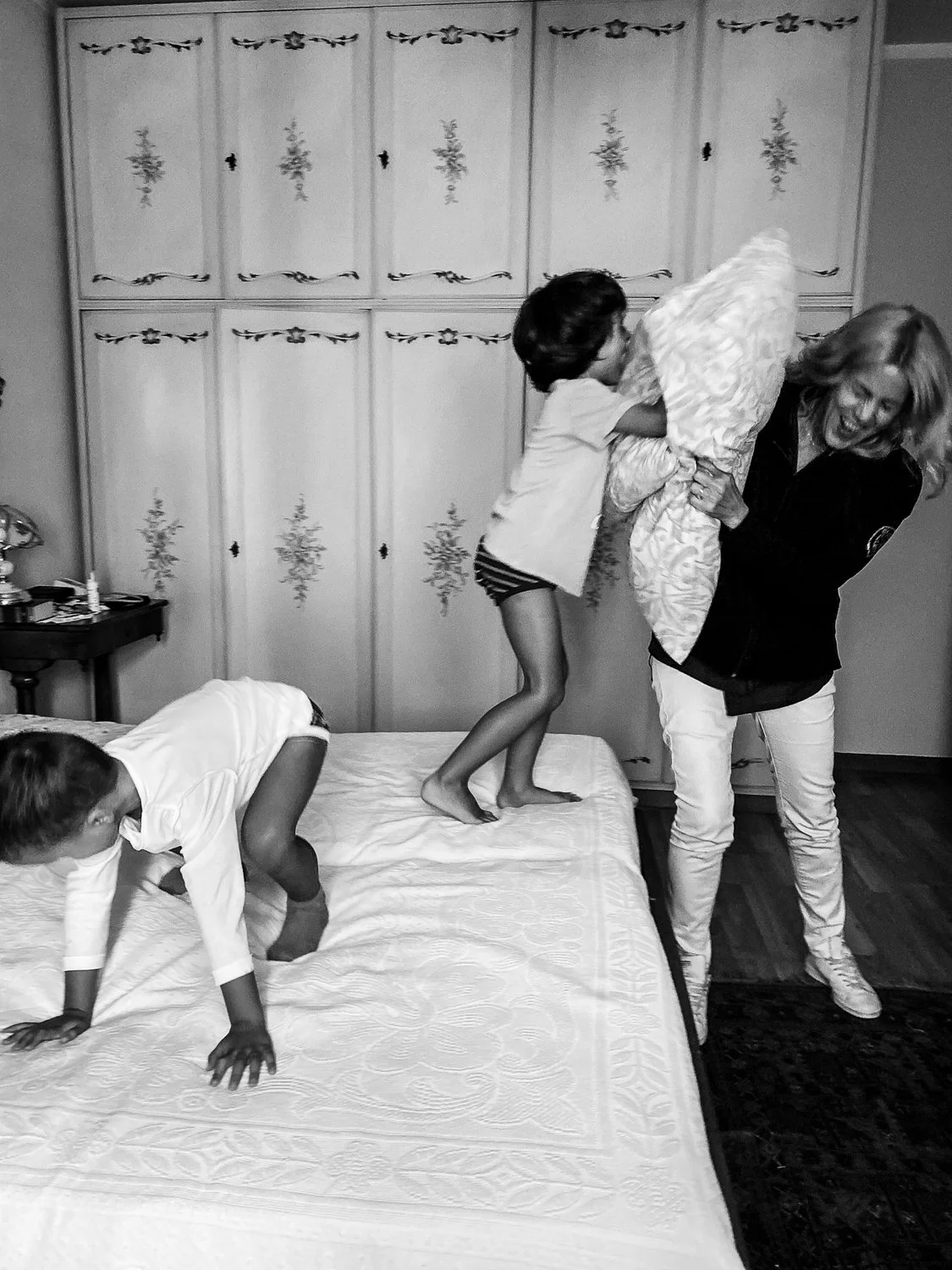 Two children and a woman playing on a bed. One child is climbing on the woman holding a pillow, another child is crawling on the bed.