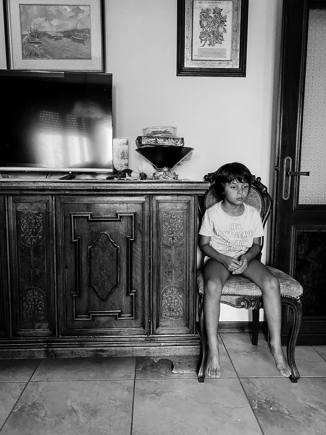 A young girl sitting on a vintage chair against a cabinet in a living room, with framed art on the wall and a TV on the cabinet.