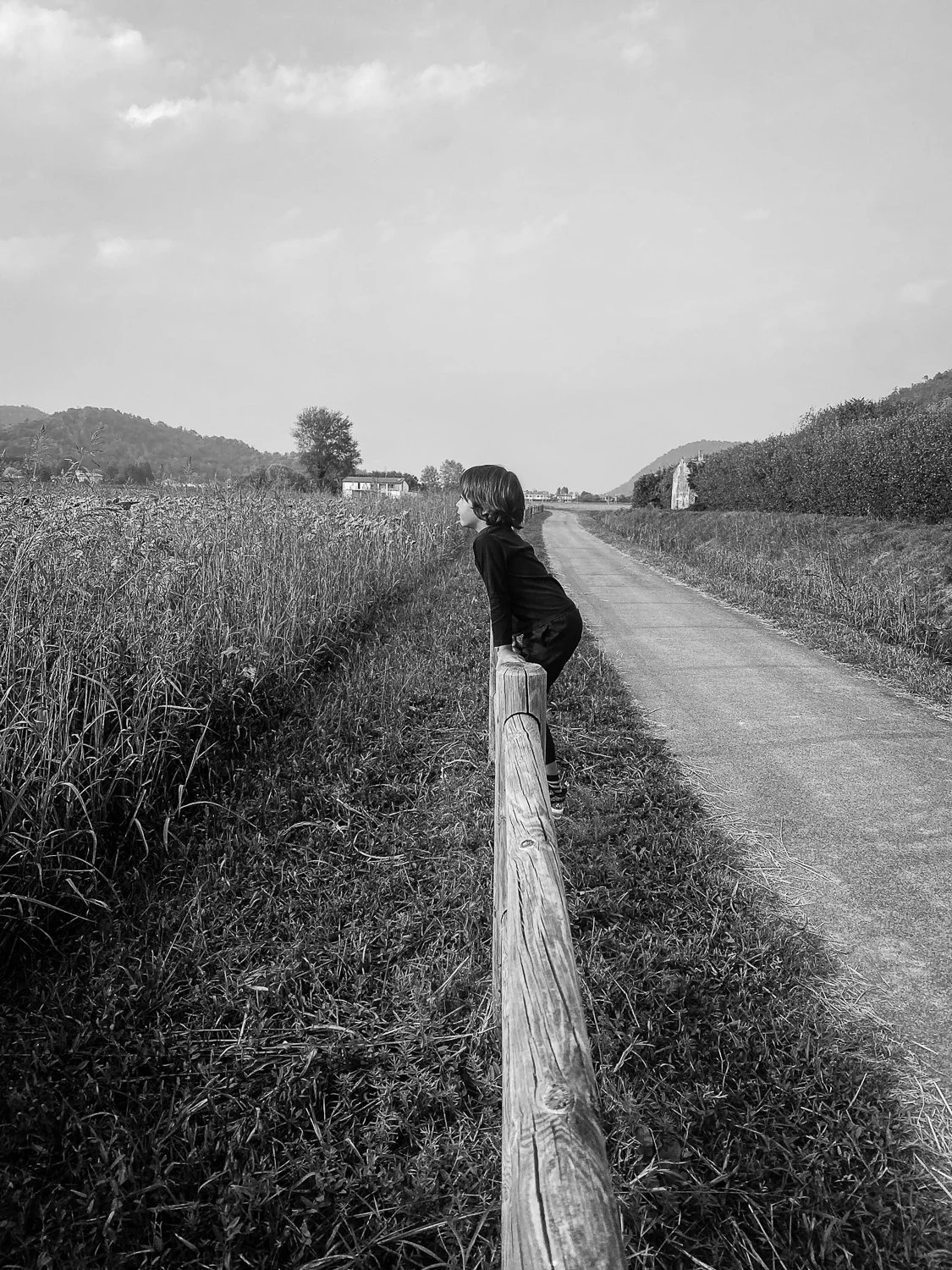 A child leaning over a wooden railing along a rural road, looking into the distance, with fields and hills in the background, in black and white