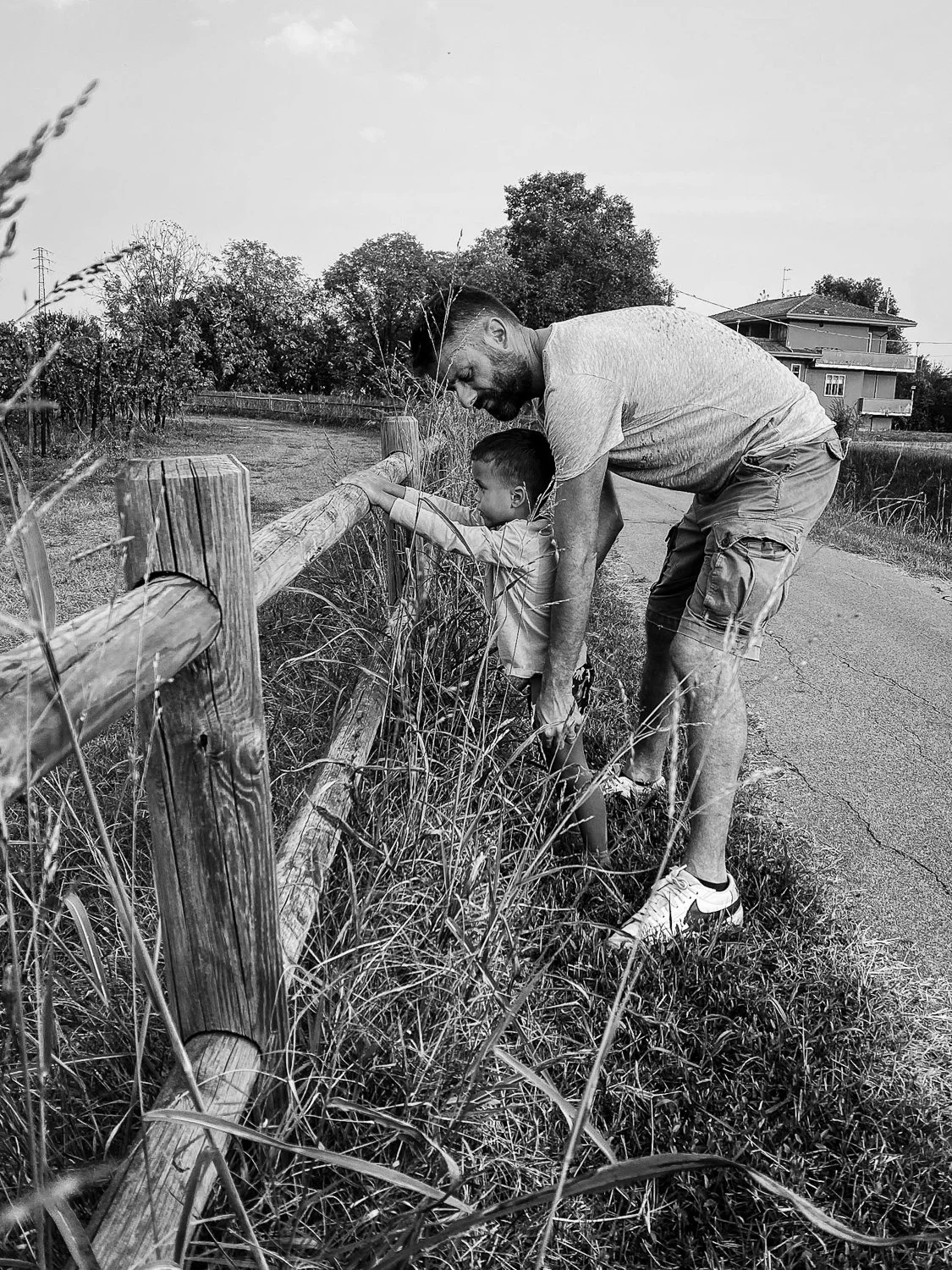 A man and a young boy are leaning over a wooden fence beside a sidewalk, looking at the grass and weeds beyond the fence. The man is assisting the boy, and both appear to be engaged in observing or exploring the outdoors. There are trees and houses i