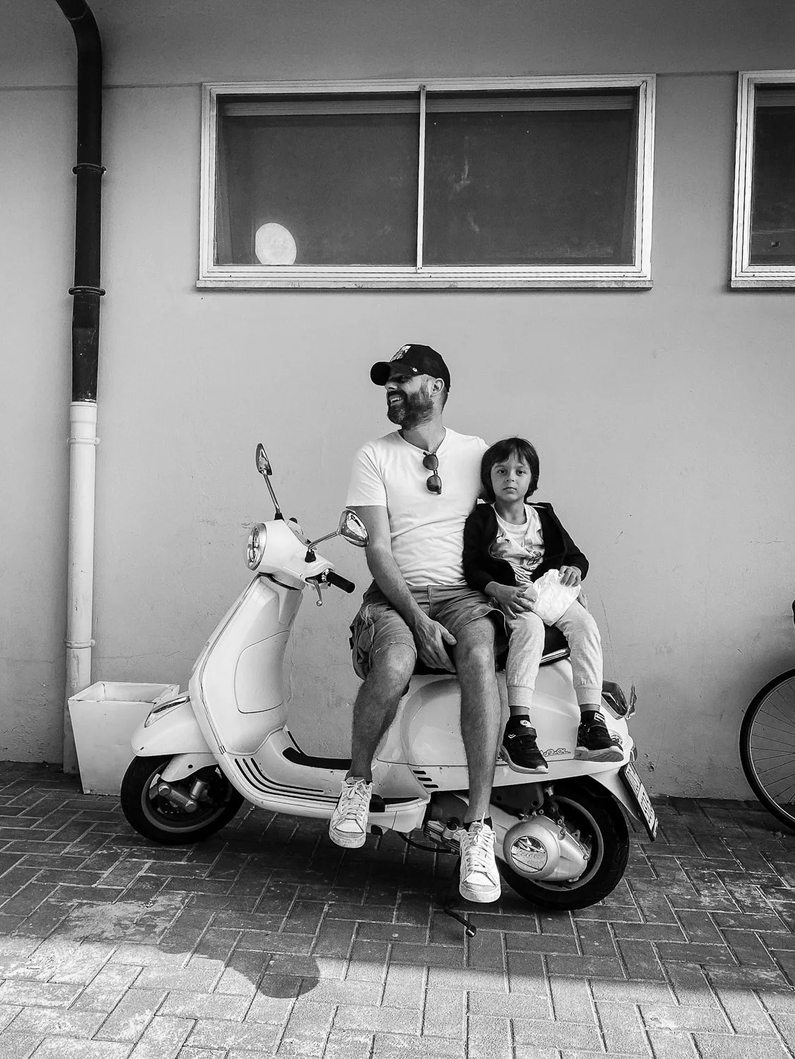A man and a young boy sitting on a white scooter parked against a plain wall with two windows and a window. The man is wearing a white t-shirt, shorts, a cap, and sunglasses hanging on his shirt, and is smiling. The boy, sitting on the man's lap, has