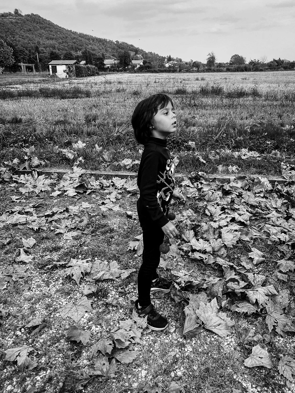 A young boy stands outdoors on a leaf-covered patch of ground, surrounded by a rural landscape with fields, houses, and hills in the background. The image is in black and white.