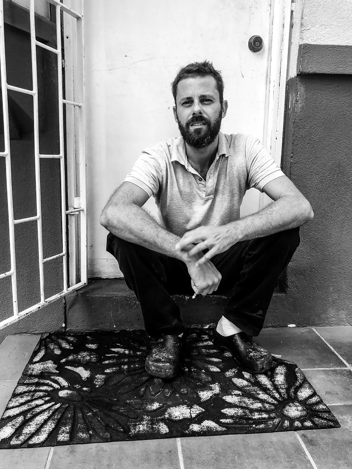 A man with a beard and short hair squatting on a decorative outdoor mat, wearing a light-colored polo shirt, dark pants, and clogs, outside a door with a metal security grill and a wall with a light and dark paint color.
