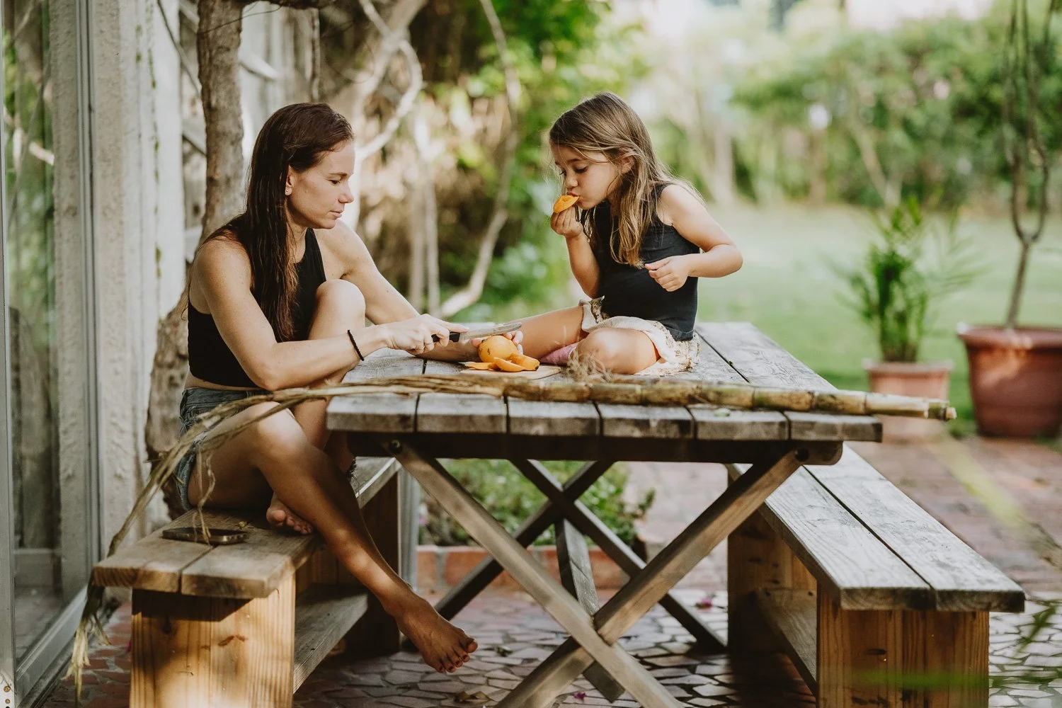 A woman and a young girl sitting at a wooden picnic table outdoors, the woman looking at her phone and the girl eating a snack.