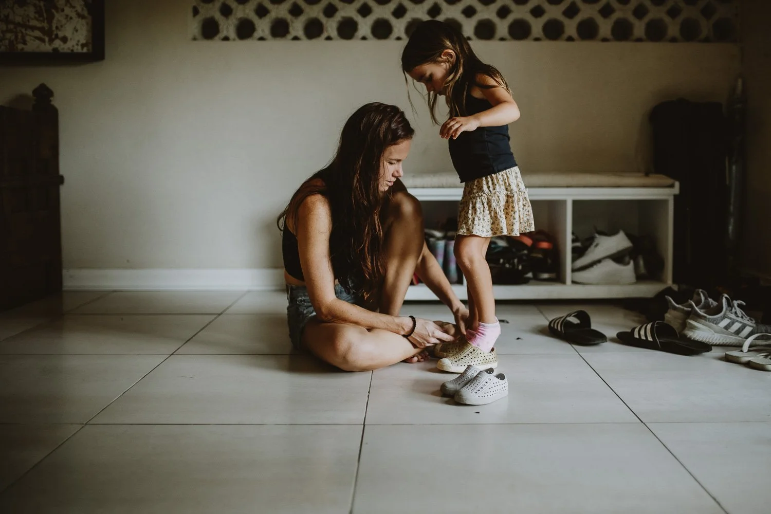 A woman helping a young girl put on her shoes inside a room with tiled floors and a white shelf in the background.