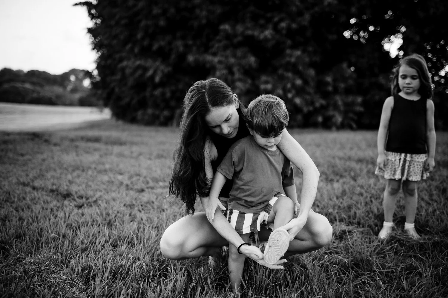 A woman helping a young boy put on his shoe in a grassy field with two other children watching nearby.
