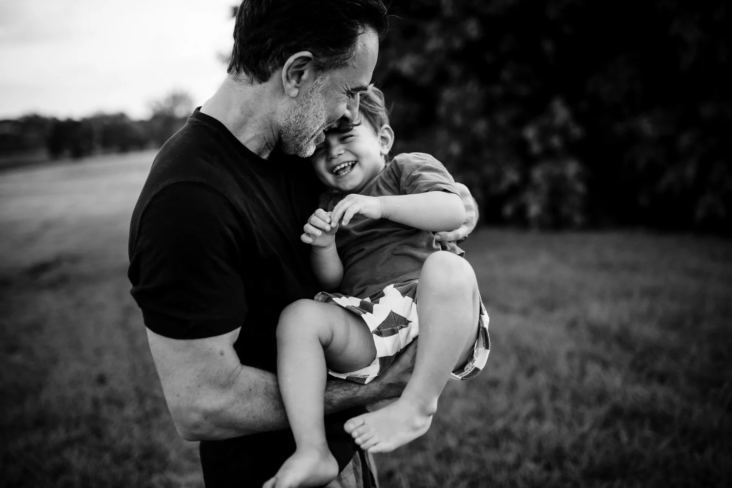 A man holding a laughing young boy outdoors, both smiling and close together in a black and white photograph.
