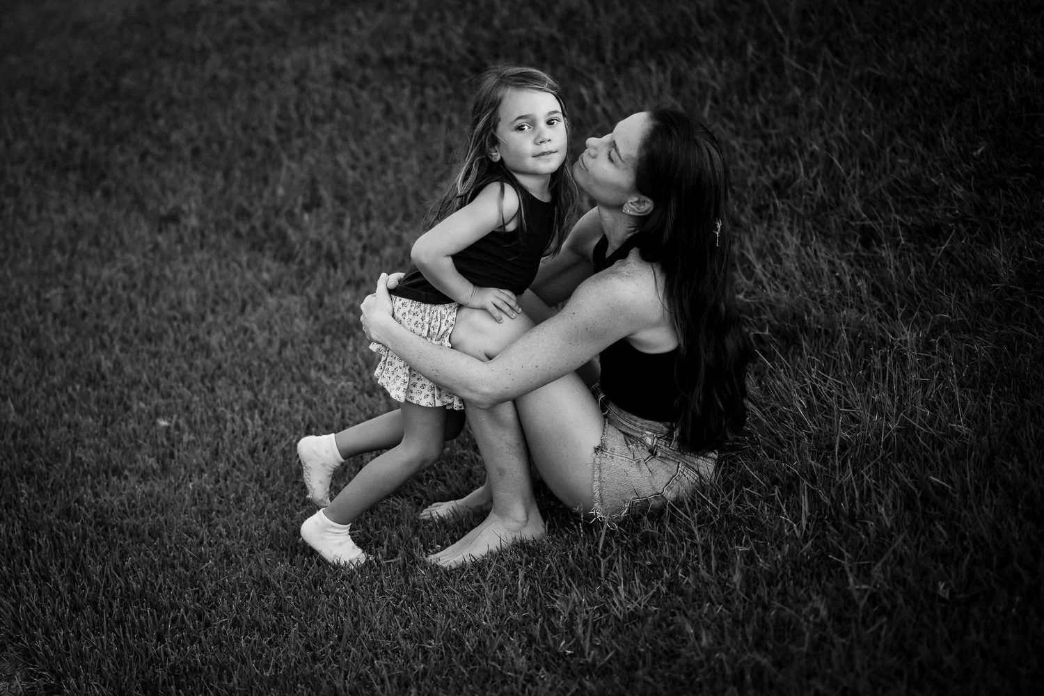 A woman and a young girl sitting on grass, sharing a close moment; the woman kisses the girl on the cheek while the girl looks at the camera.
