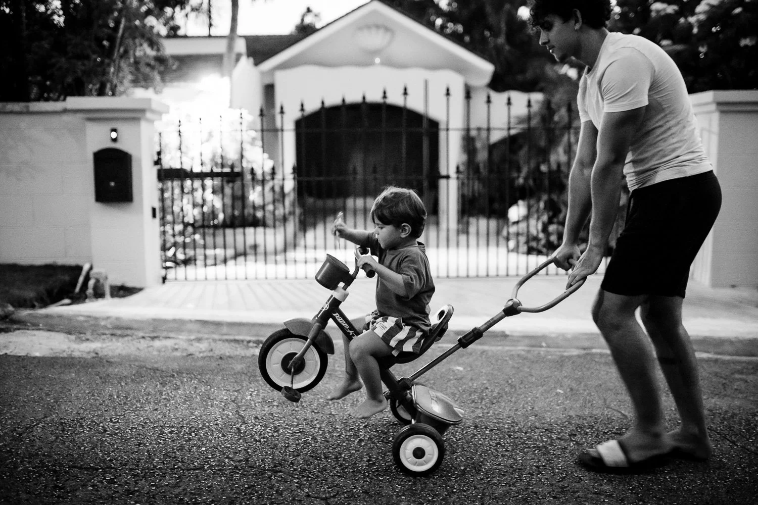 A young child riding a small bike with training wheels as an adult pushes him from behind on a residential street, with a house and gate in the background.